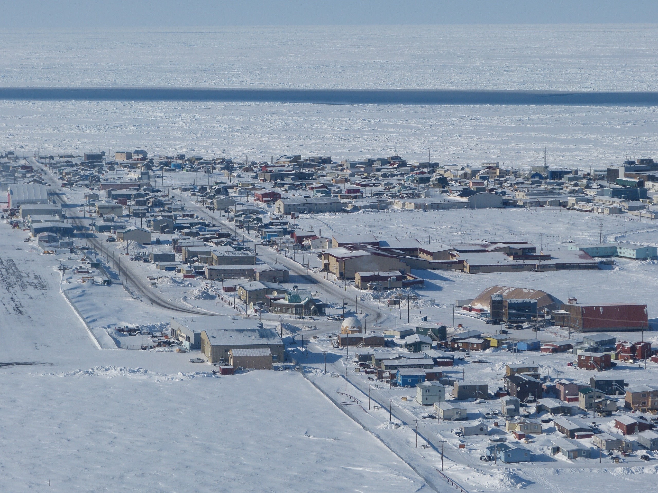 Aerial view of Utqiaġvik, Alaska, showing a snow-covered coastal town bordered by sea ice. A long, narrow strip of open water called an open flaw lead cuts across the frozen ocean, separating the solid landfast ice attached to shore from the drifting pack ice farther offshore.