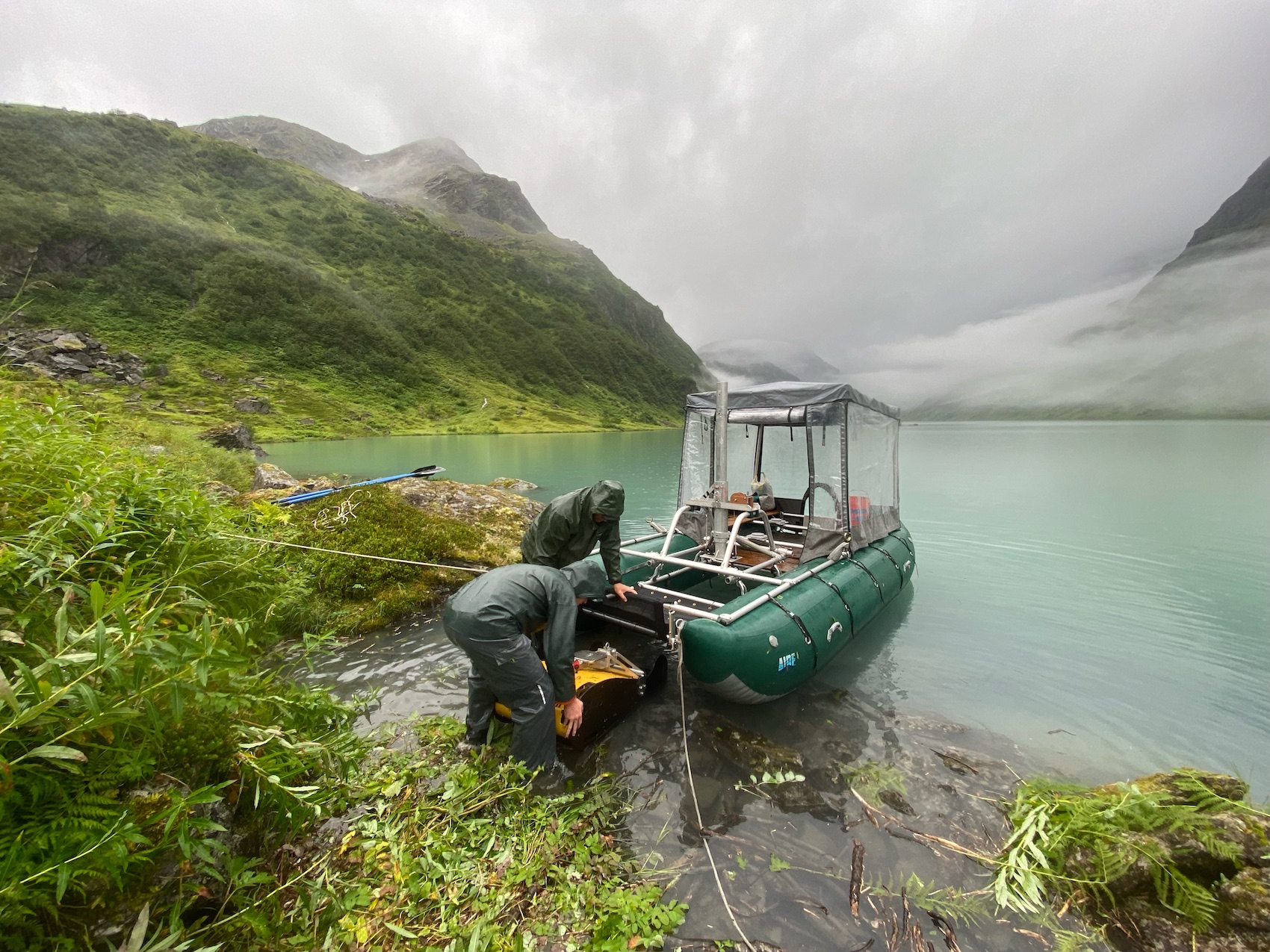 Two people work next to a pontoon boat floating on a lake surrounded by green alpine tundra and mountains obscured in foggy clouds.