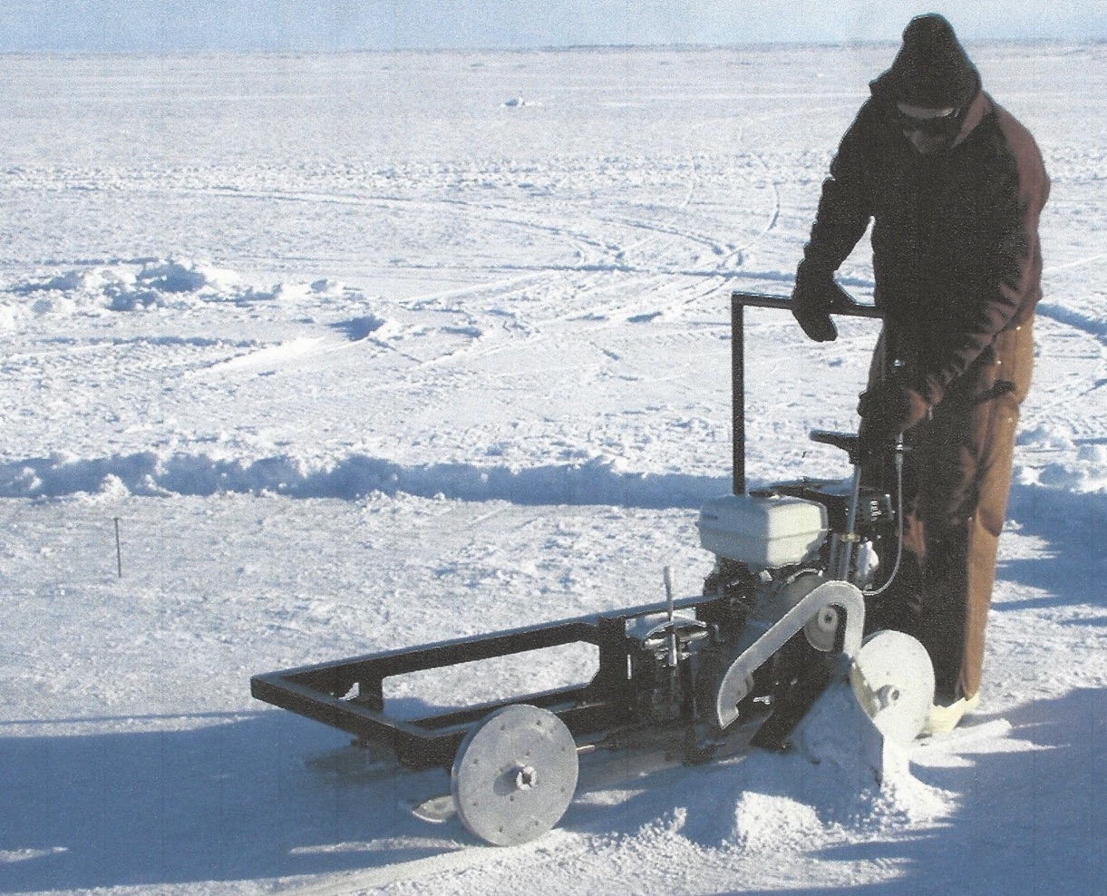 Standing on snow-covered sea ice, a man in coveralls operates controls on a gas-powered saw mounted on a steel-framed sled.