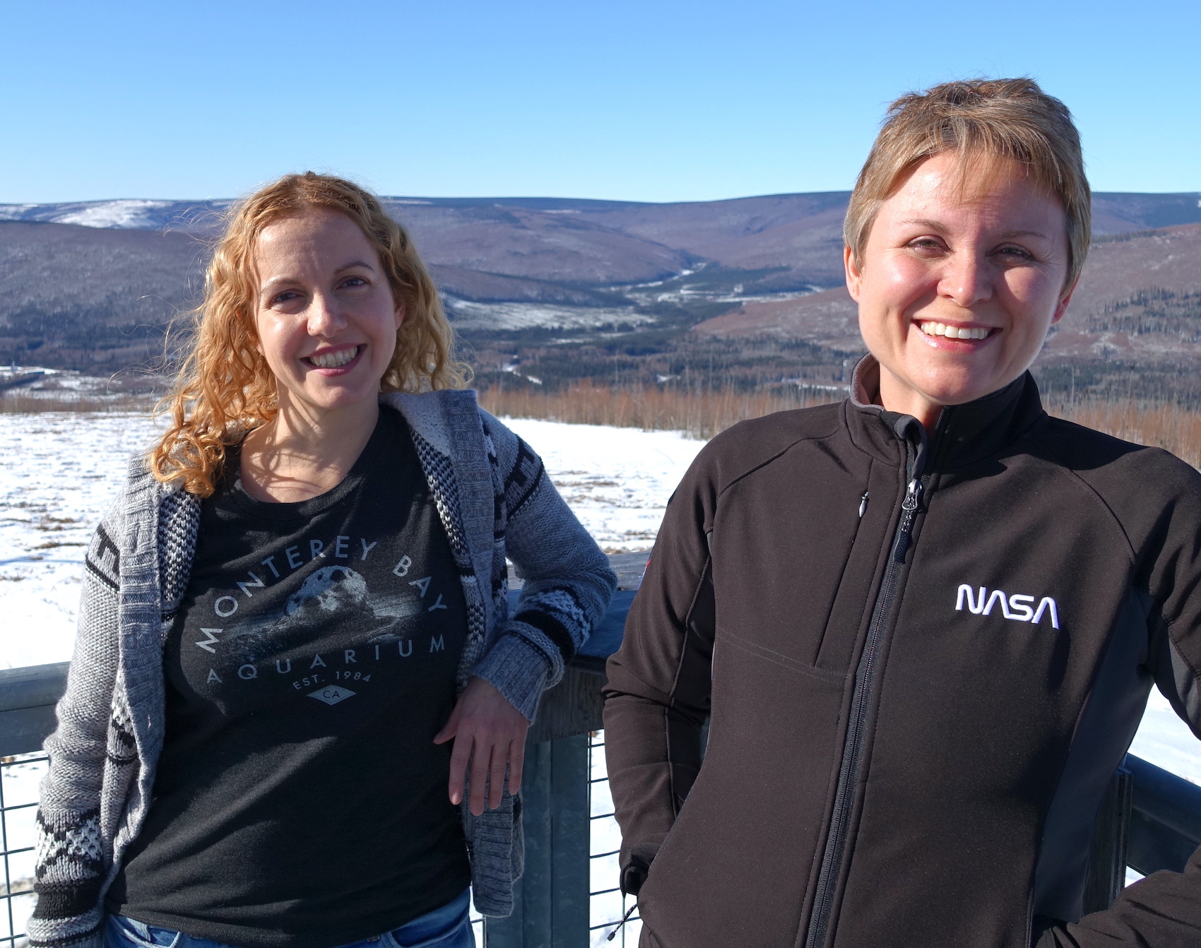 Two smiling women lean on an outdoor deck railing with a snowy field and rolling hills in the background. 