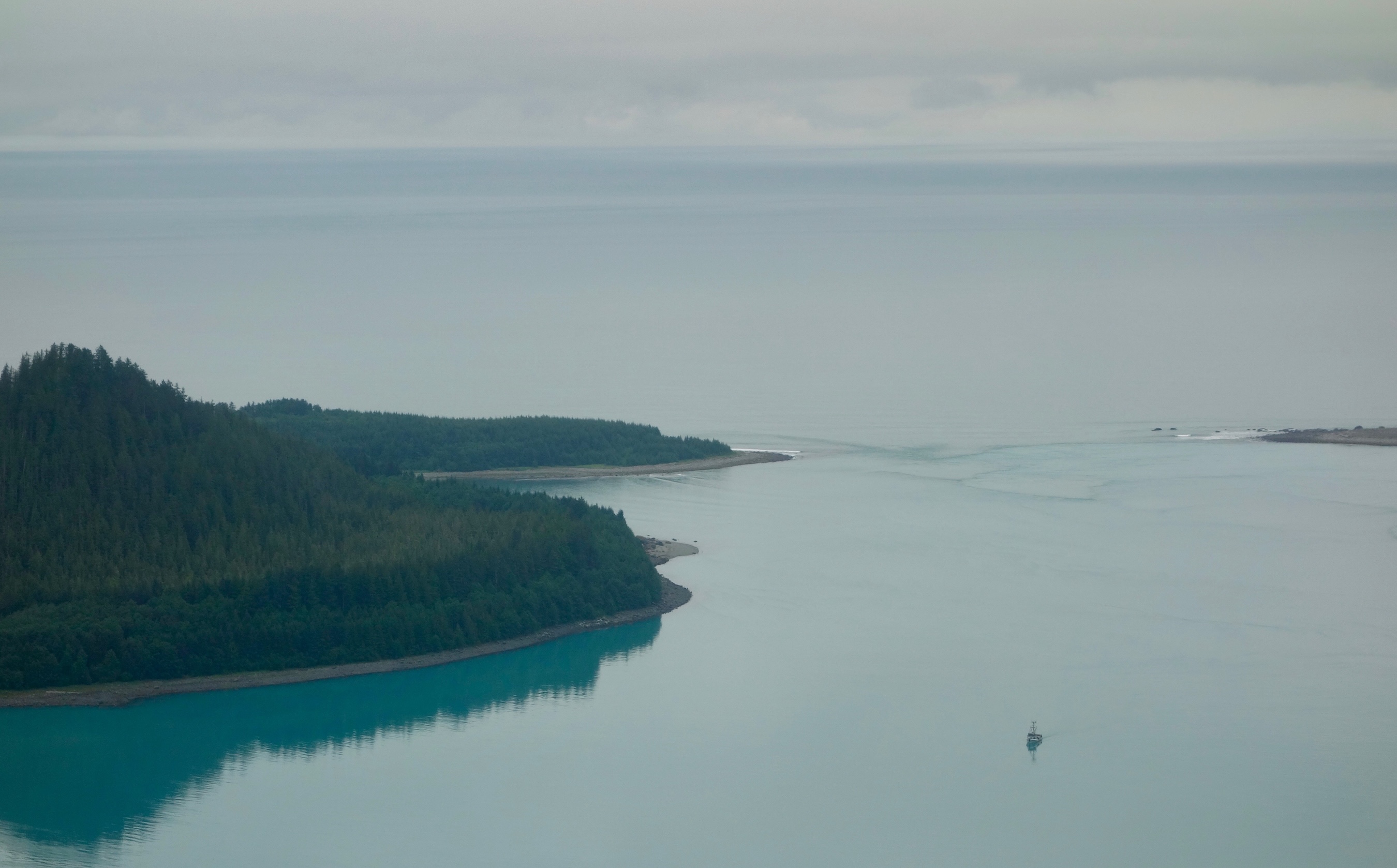 A boat sits in a calm bay, viewed from an aircraft. At the mouth of the bay, where it meets the ocean beyond, waves created by tidal currents are visible.