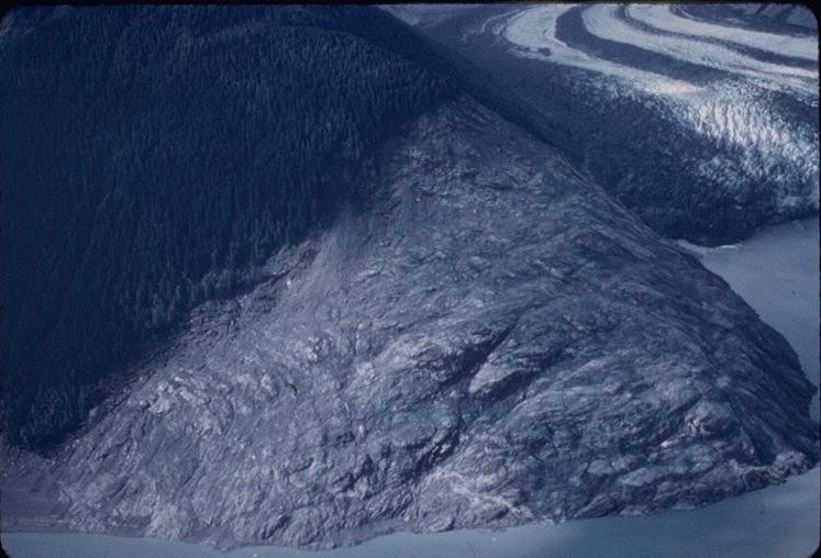 A rocky headland, stripped bare of vegetation high on its slope, rises from a body of water. In the background, a glacier terminates in the water.