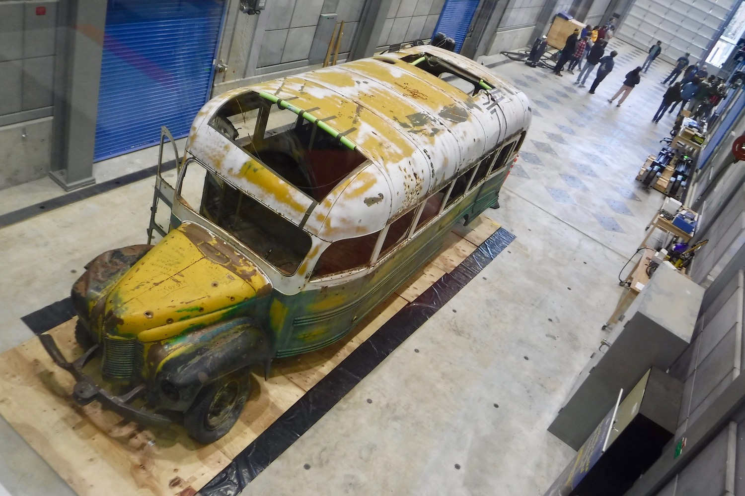 A weatherworn bus sits on a concrete floor in an elongated shop space with several overhead doors on its walls.