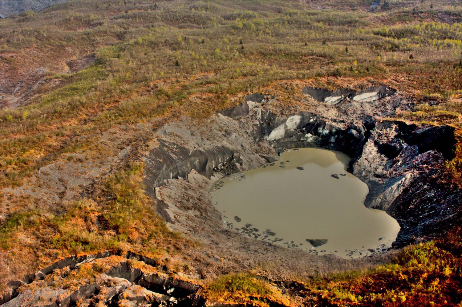 A hole in a glacier, a few hundred yards wide and lined with ice and gravel, is surrounded by a low forest in fall colors.