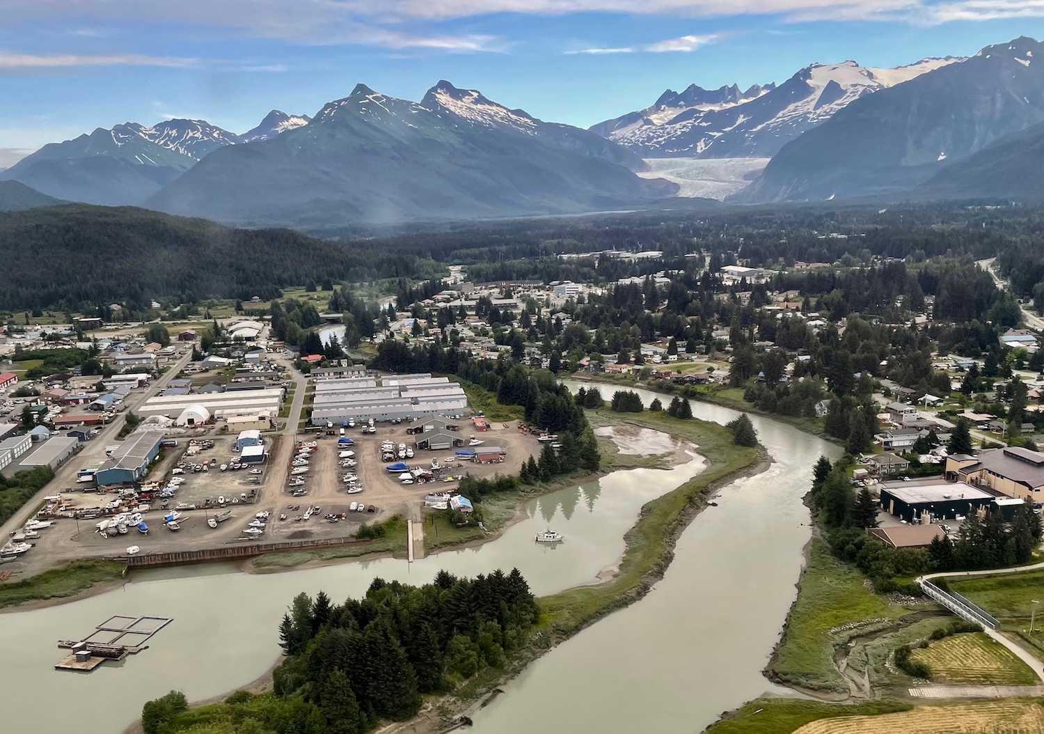 Homes and businesses sit along a silty river with mountains and a glacier in the background.