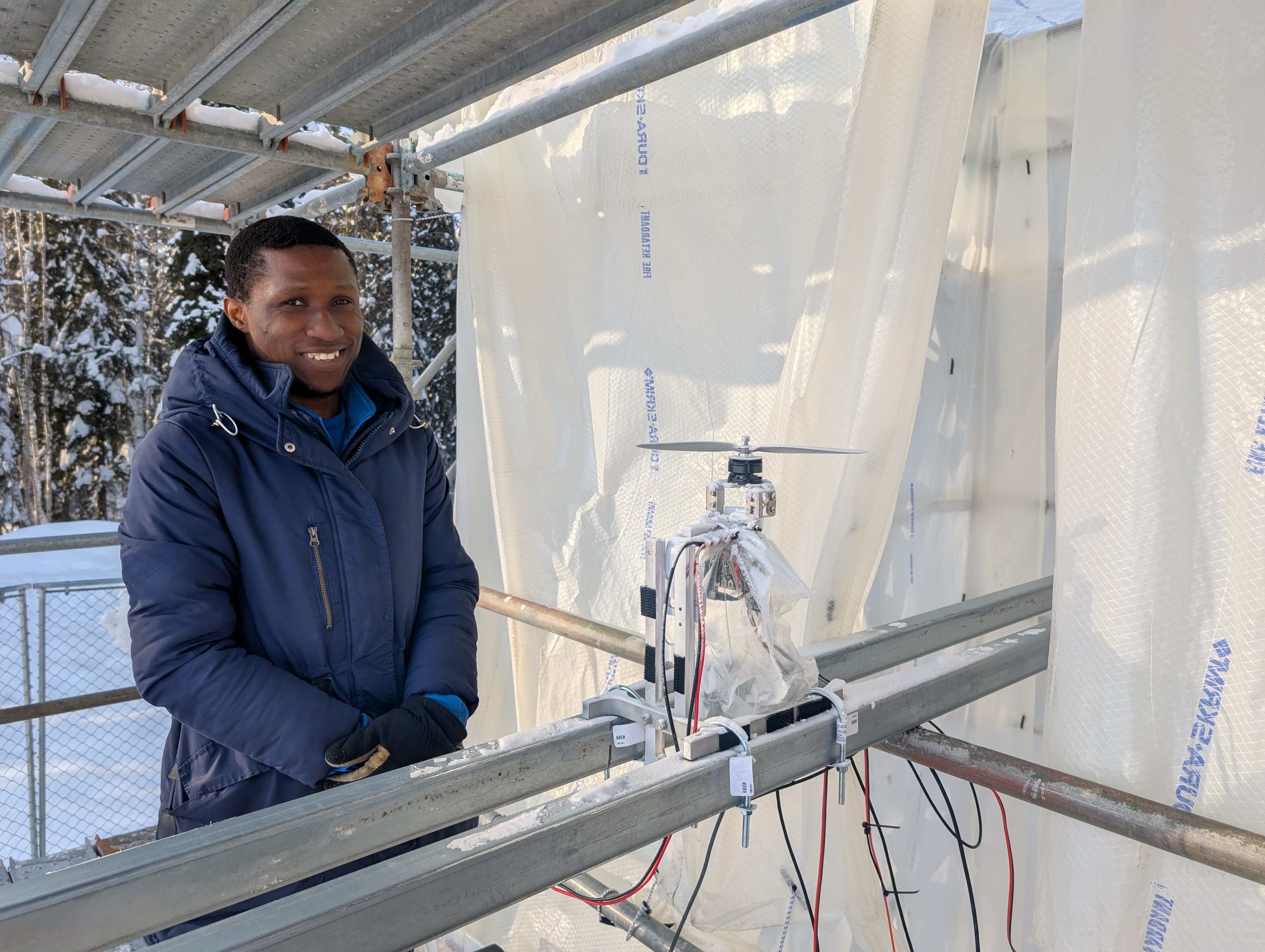 Doctoral student Morakinyo Labiran stands at ACUASI’s ice tower beside his planned test item, a small rotor assembly mounted on metal rails and surrounded by wiring. He wears a heavy blue winter coat and gloves inside the translucent plastic-lined, scaffold-supported enclosure.