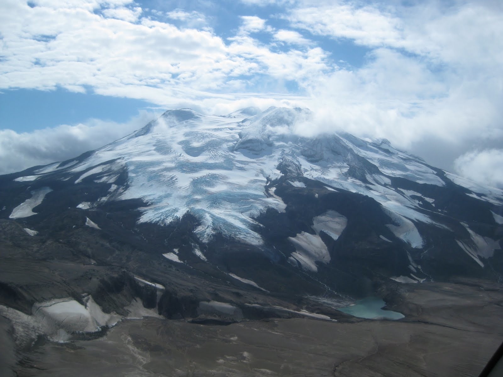 A glacier-capped volcano rises through broken clouds and sunshine. A blue lake sits at the base of the volcano.