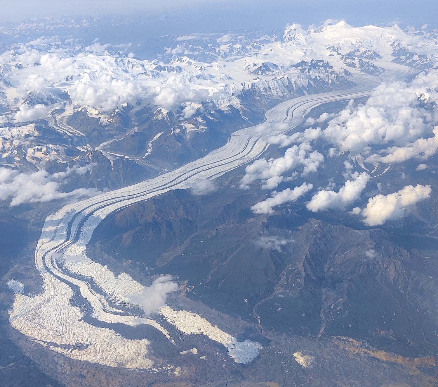As viewed from an aircraft, a vast glacier winds through a mountainous valley, starting at high snow-covered peaks in the background and ending in a splayed terminus among snow-free peaks in the foreground.