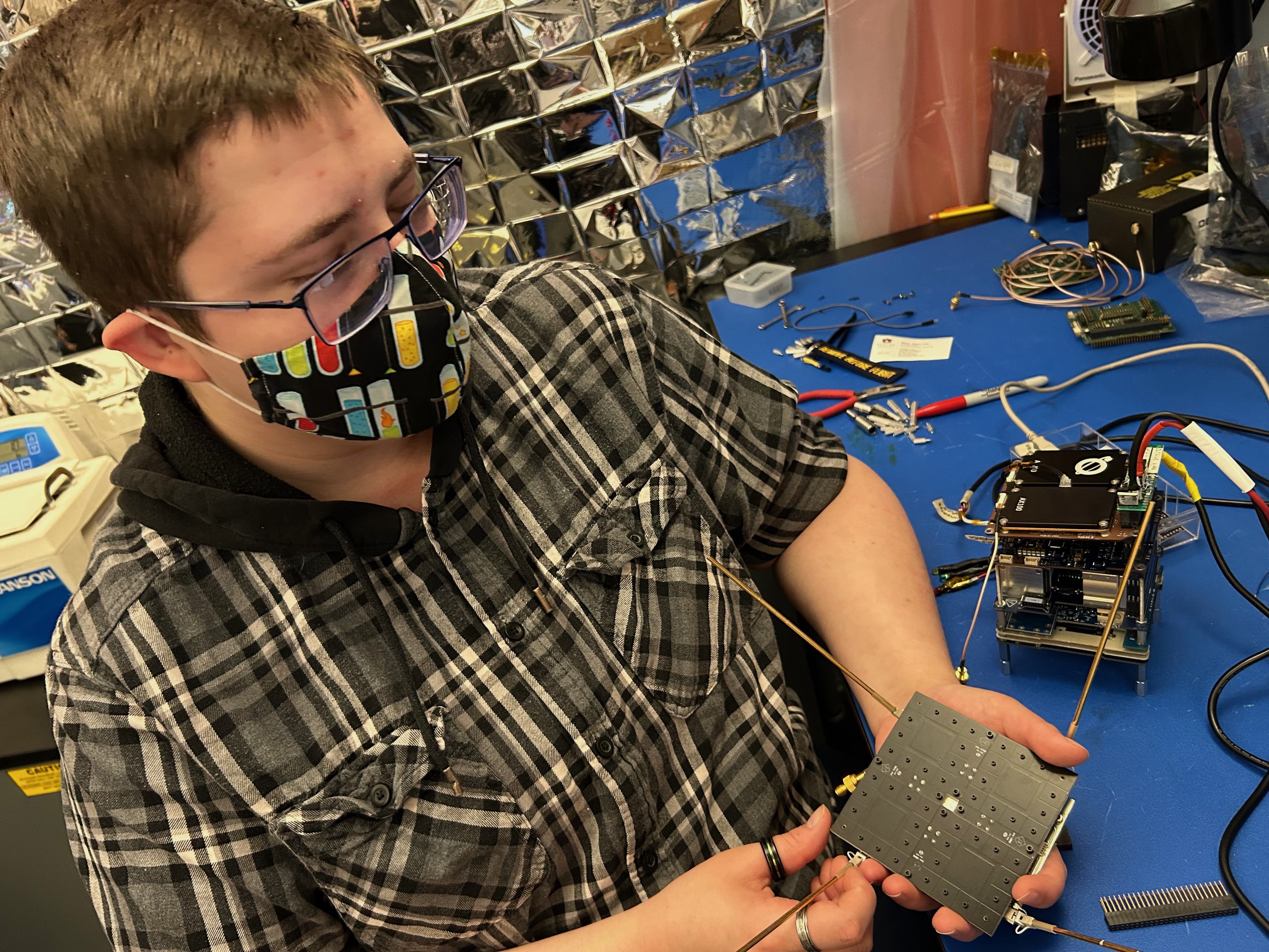 Student Caleb Fronek, the nanosat project manager, holds the retrodirective and UHF antennas. The avionics test stack is on the tabletop.
