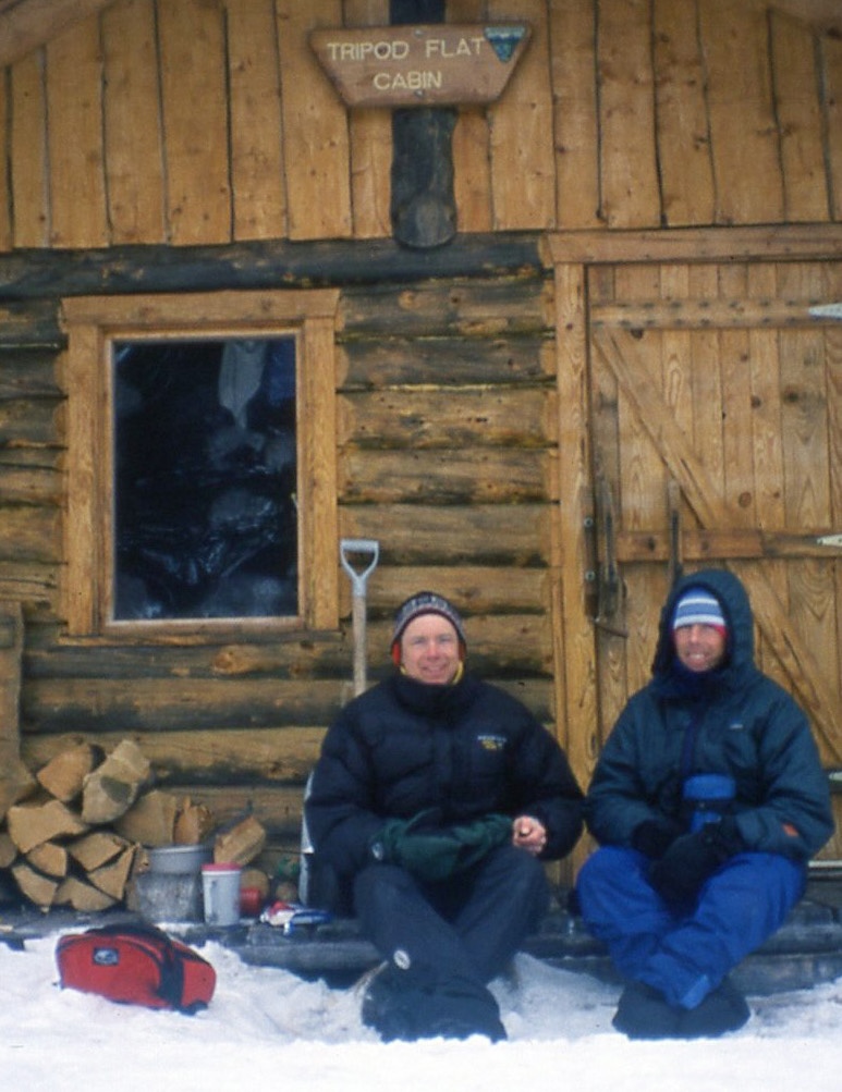 Two men in warm winter gear sit outside a log cabin with their feet resting in snow. Cooking gear, firewood and a snow shovel sit on the porch next to them. A red gear bag sit on the snow.