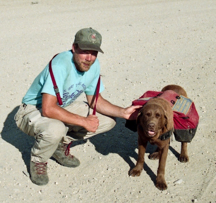A blond bearded man in a blue T-shirt and green ball cap, holding a leash over his shoulders, squats next to a brown Labrador retriever wearing a red dog pack. 