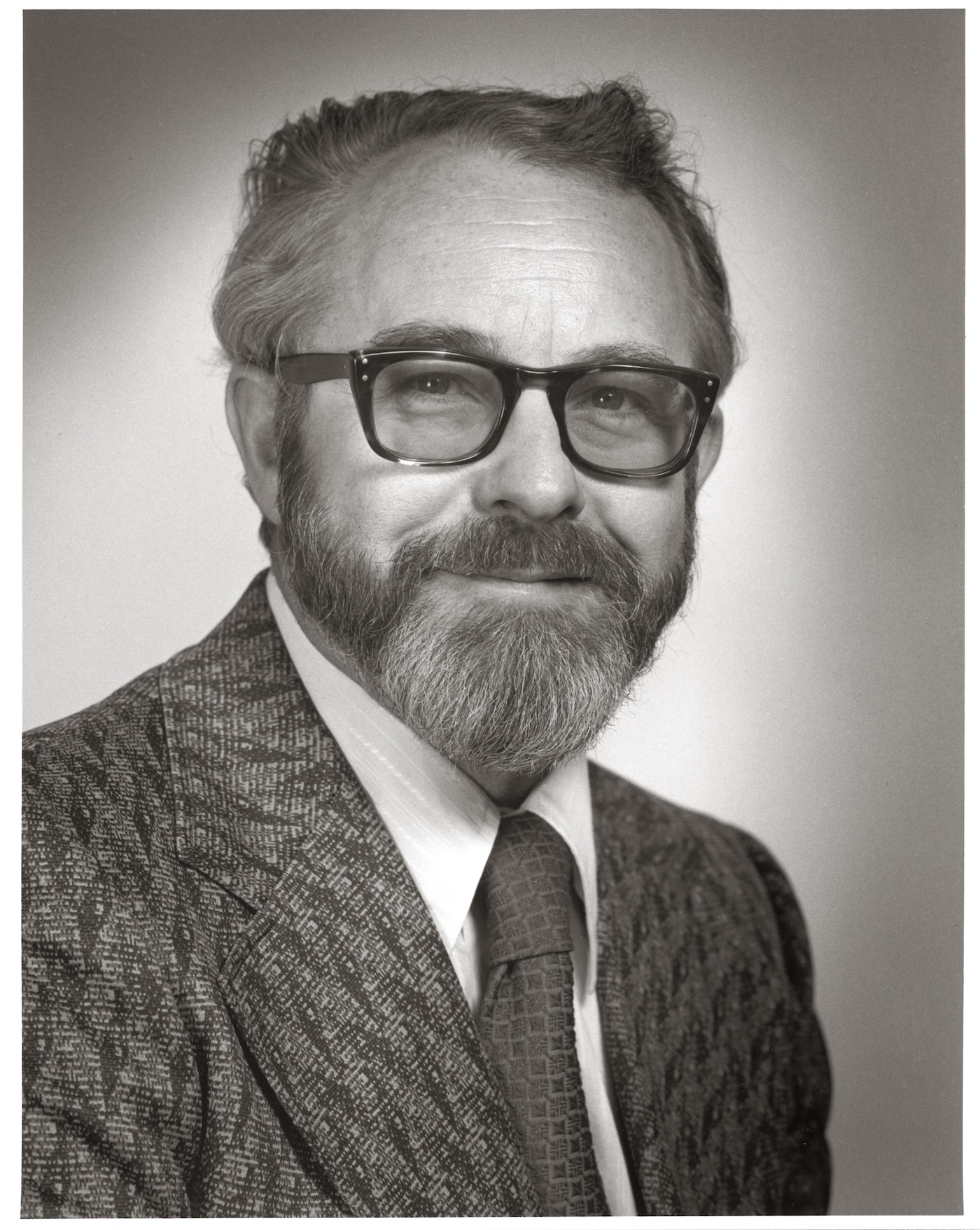 A bearded man with glasses poses for a portrait while wearing a jacket and tie.