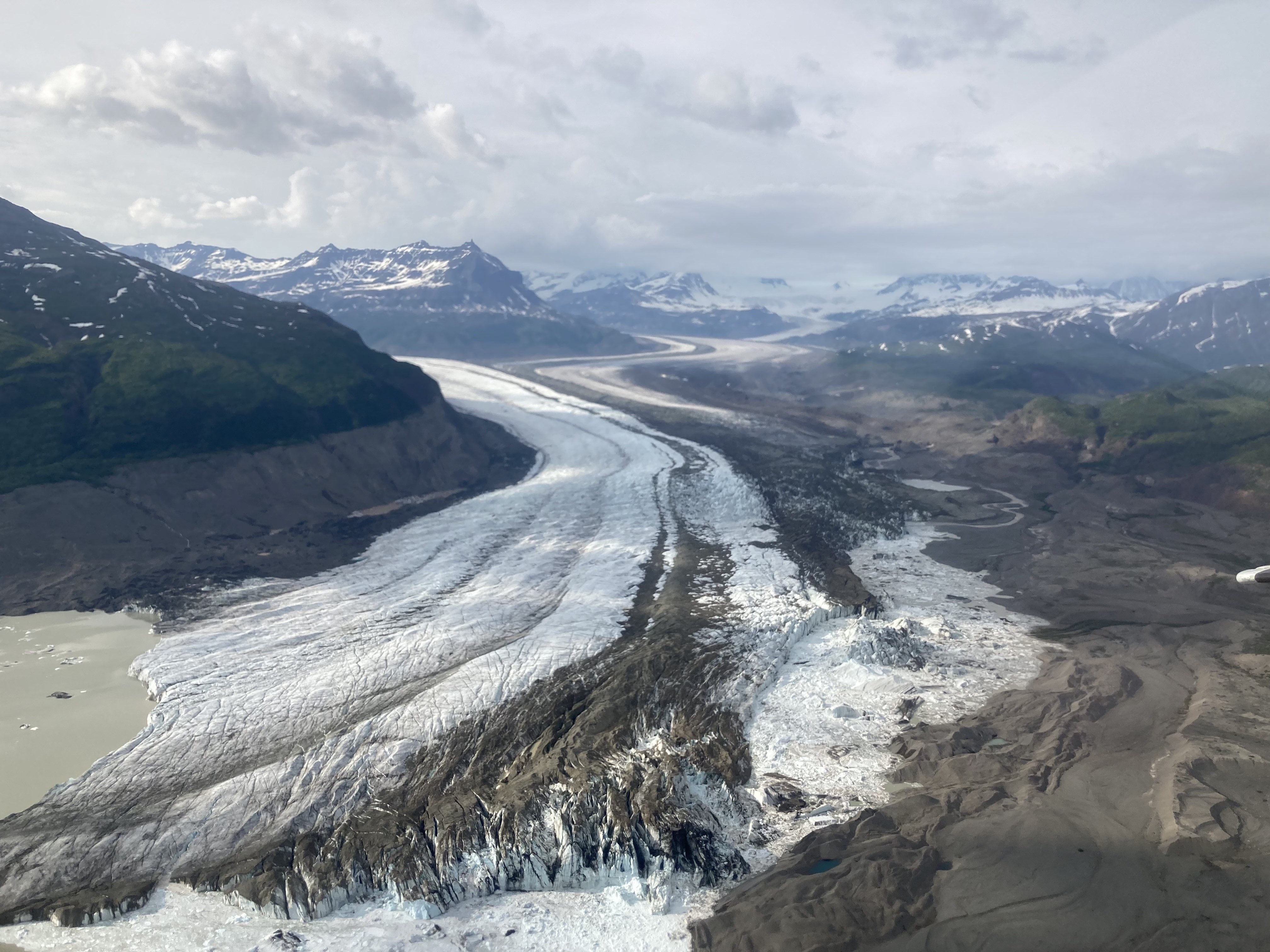 A sweeping aerial view of Nizina Glacier in summer 2022 shows a broad river of ice winding through a mountain valley 20 miles northeast of McCarthy, Alaska. Dark bands of rocky debris stripe the glacier’s surface as it curves between steep green slopes and snow-capped peaks, with braided meltwater channels spreading across the gravelly valley floor below.