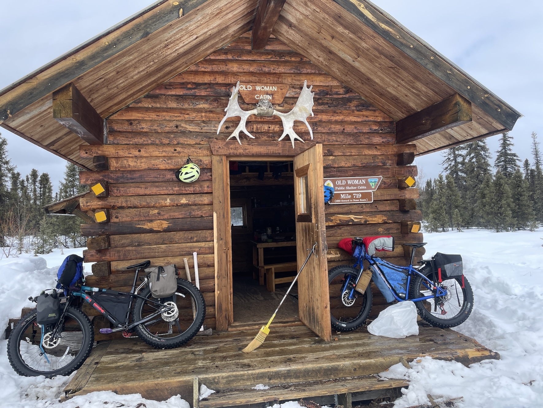 Two fat-tired bikes loaded with gear sit on the deck outside a log cabin with the door propped open with a broom. Above the door hangs a set of moose antlers and a cabin name sign. Snow surrounds the cabin, and spruce trees rise behind it. Reflector squares are affixed to four log ends. A yellow-green bike helmet hangs on the exterior wall.