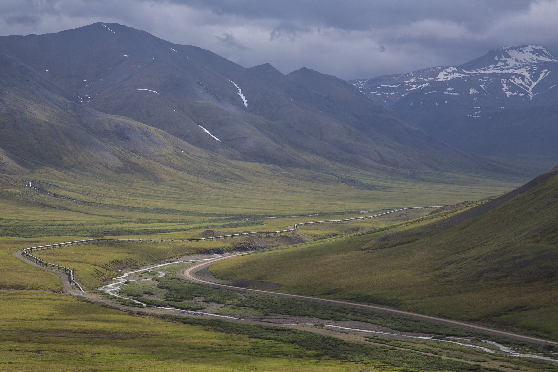 A road, river and pipeline parallel one another along the bottom of a green tundra-covered mountainous valley.