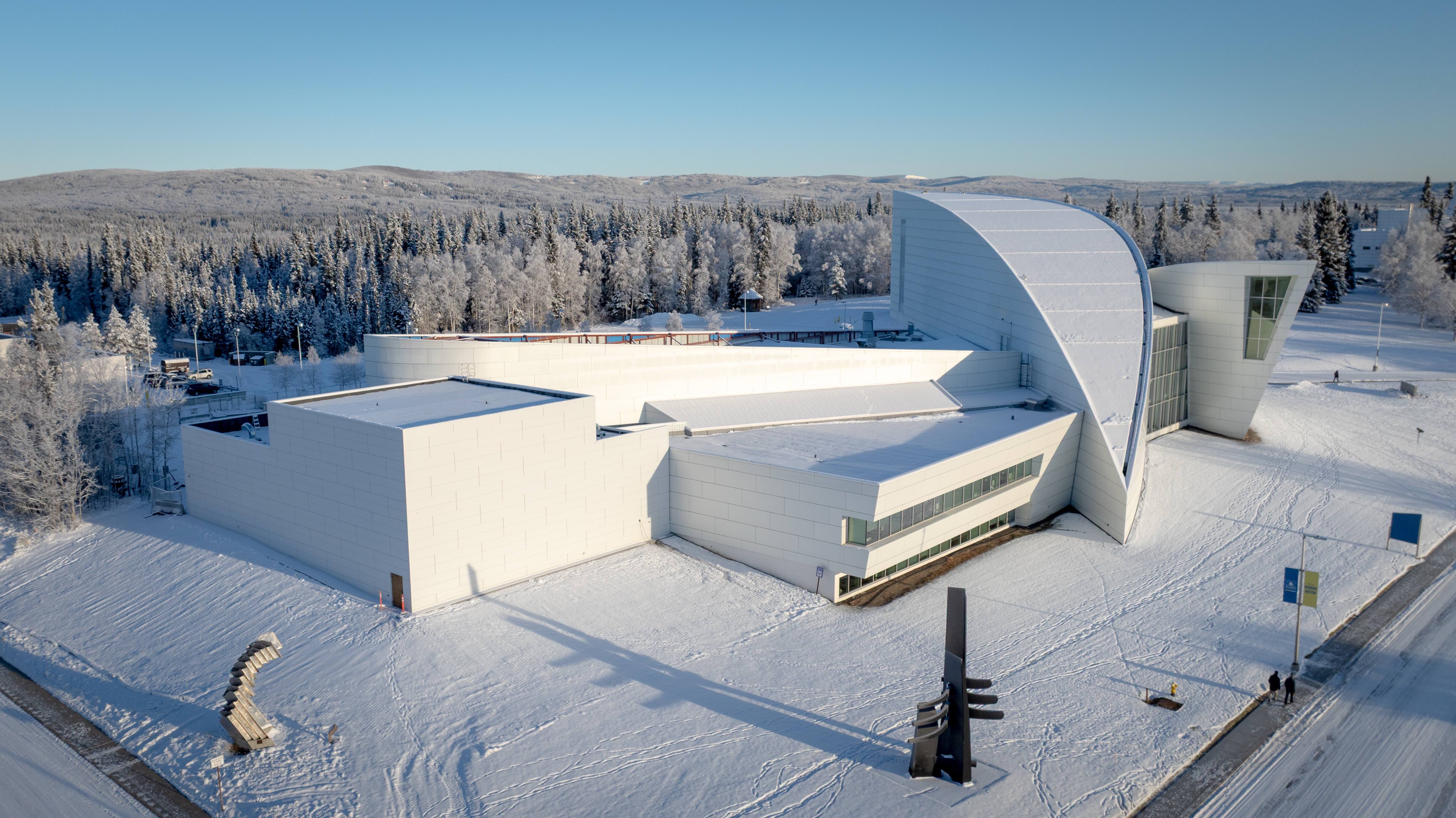 Aerial view of the Walt and Marita Babula Planetarium shows a white, modern complex surrounded by snow-covered forest and hills under clear winter light. The image, taken in November 2025, shows the planetarium addition to the west side of the UA Museum of the North. The square planetarium addition is visible at left.