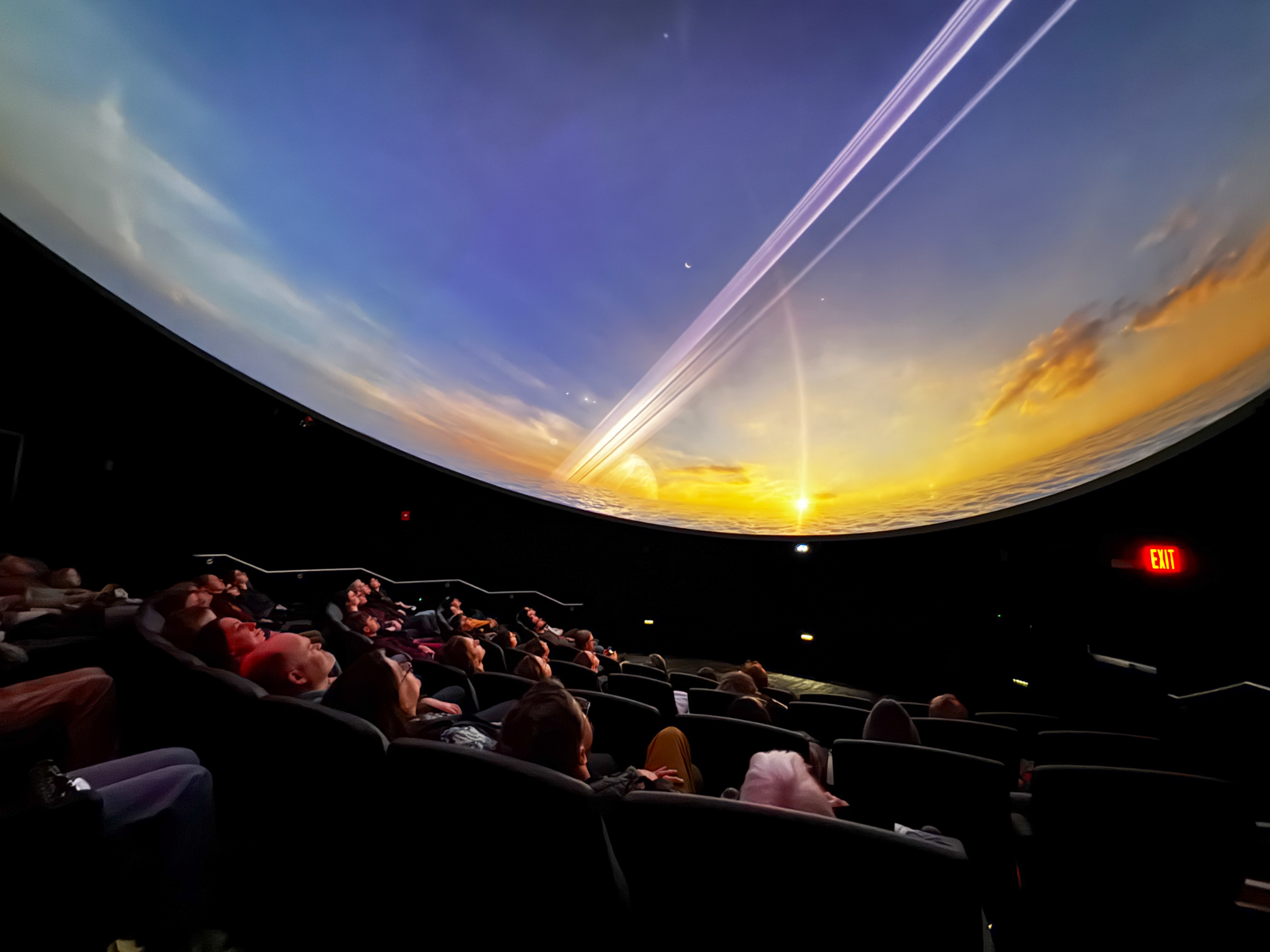 Audience members sit in reclined theater seats, looking up at a curved dome screen displaying a vivid space scene during a movie screening. The image shows the interior of the Walt and Marita Babula Planetarium, where attendees and guests enjoy a pre-opening screening on March 31, with the immersive projection filling the entire ceiling.