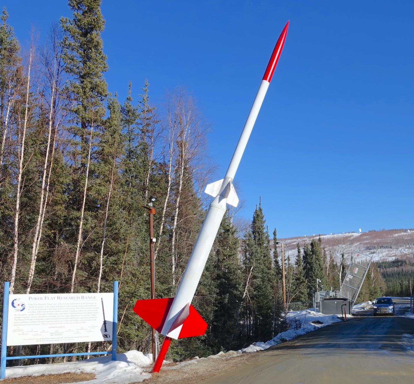 A red and white rocket leans over a road where a car drives through a raised gate. A sign sits next to the rocket. Spruce trees line the road. A hill rises in the background.