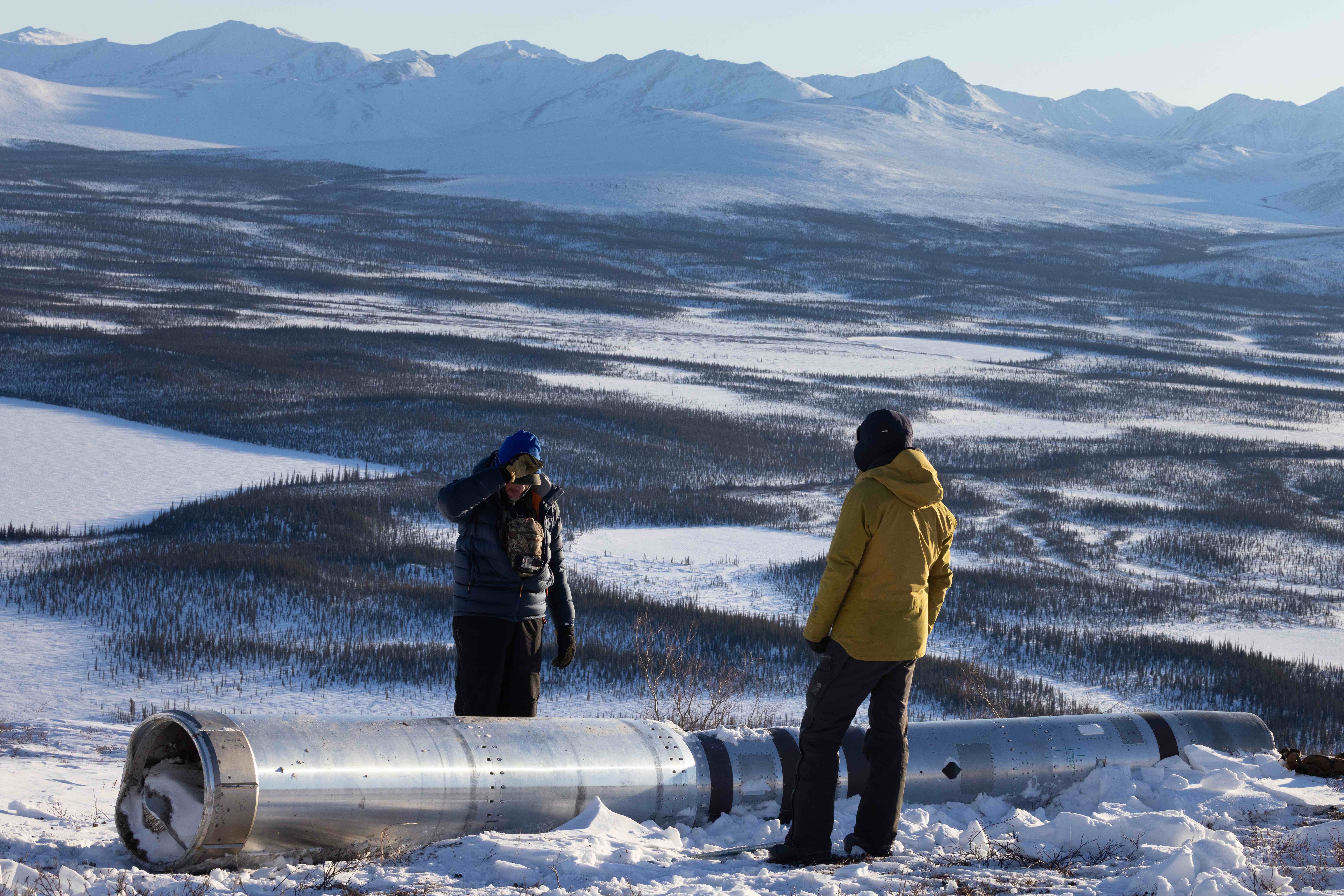Peter Elstner, left, and Brendan Lahr stand on a snowy ridge discussing the payload section of a two-stage NASA Black Brant IX sounding rocket, which lies partially buried in snow. A vast Arctic landscape stretches behind them, with rolling, snow-covered terrain and distant mountains under clear winter light.