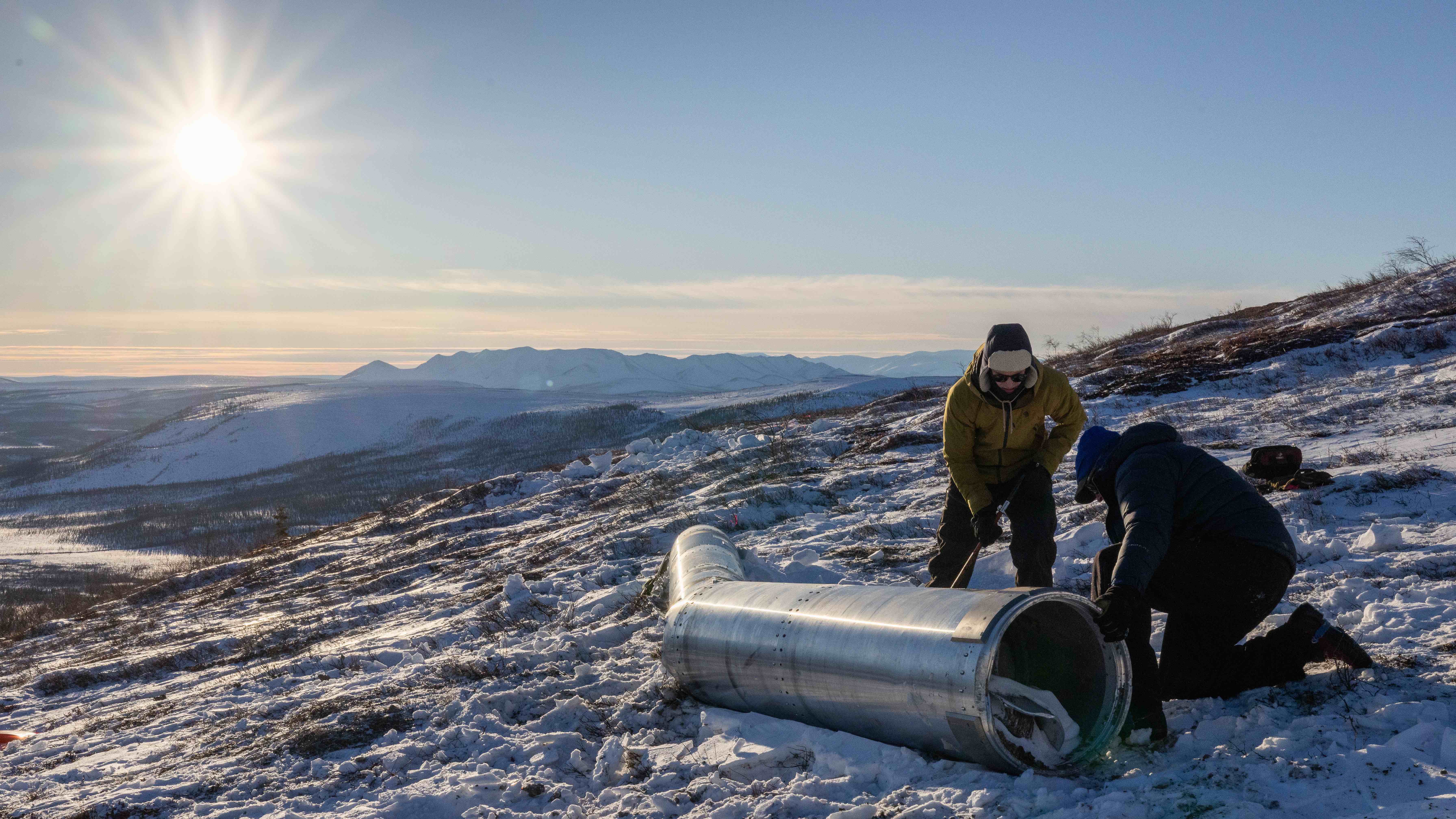 Peter Elstner, right, and contracted assistant Brendan Lahr work on a snow-covered hillside to prepare the PolarNOx payload section for its return to Poker Flat Research Range. The cylindrical rocket component lies on the ground between them as they handle tools, with low winter sunlight and distant mountains stretching across the Arctic landscape in the background.