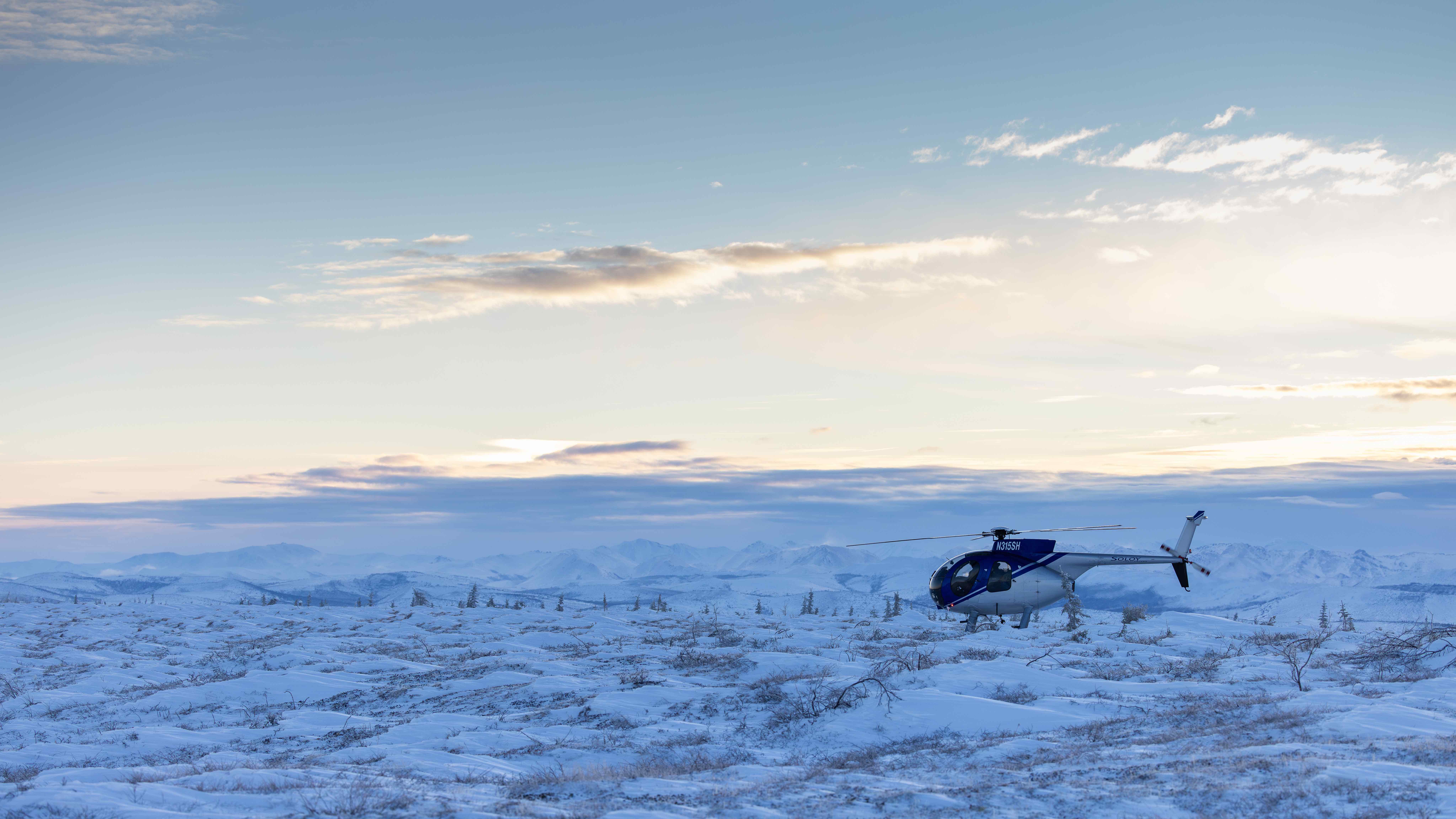 One of two helicopters involved in the PolarNOx payload recovery in early February 2026 sits on the frozen ground near the recovery site. The aircraft rests on a snow-covered tundra with low Arctic vegetation, while distant mountain ranges and a pale winter sky stretch across the background.