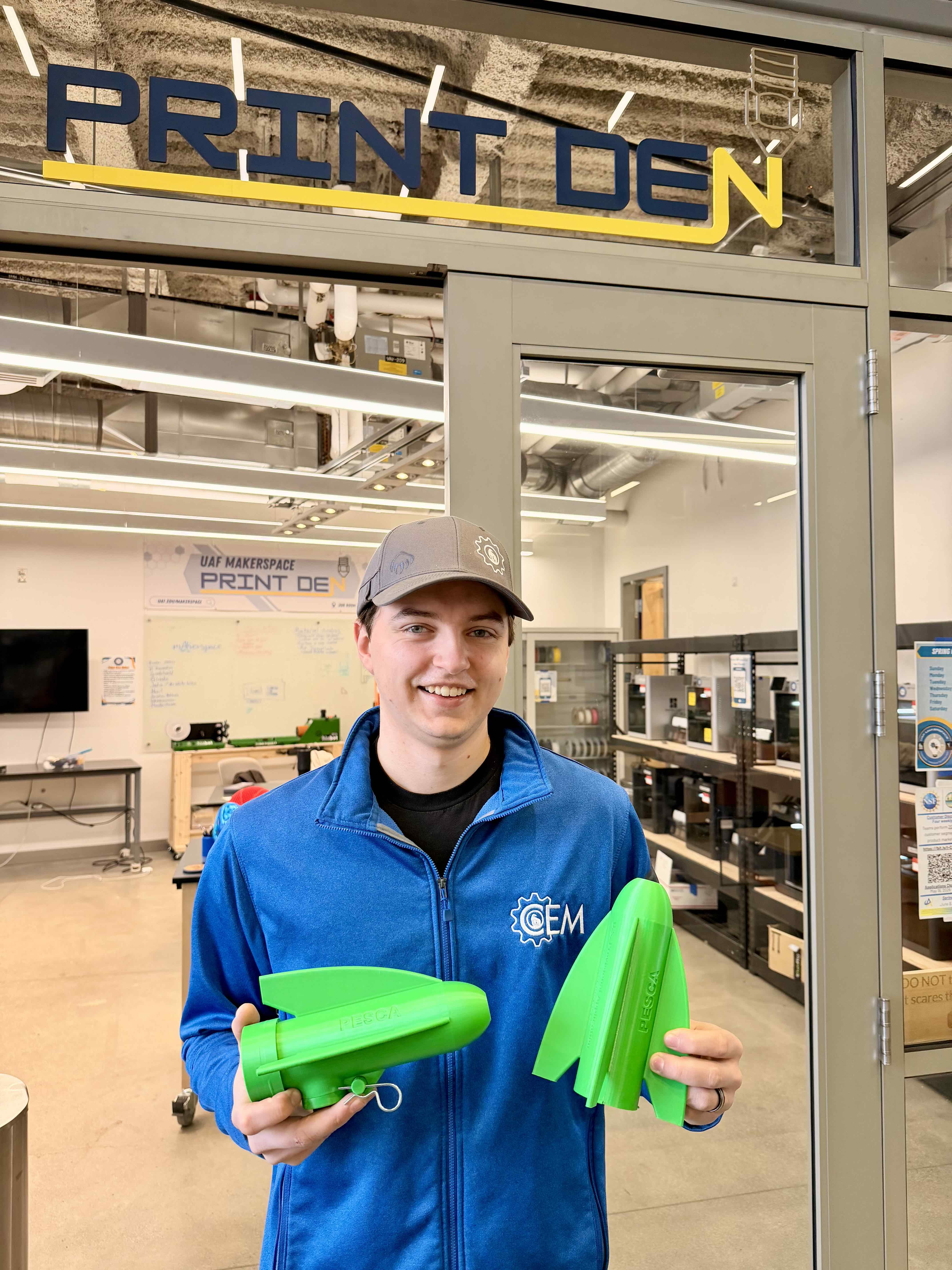 A man stands in front of a glass-walled makerspace labeled “PRINT DEN.” He is wearing a gray cap and a blue jacket with a CEM logo and is holding two bright green 3D-printed components—one assembled and one in separate fin-like pieces. Behind him are shelves of 3D printers, worktables, and a whiteboard inside the UAF Makerspace lab.