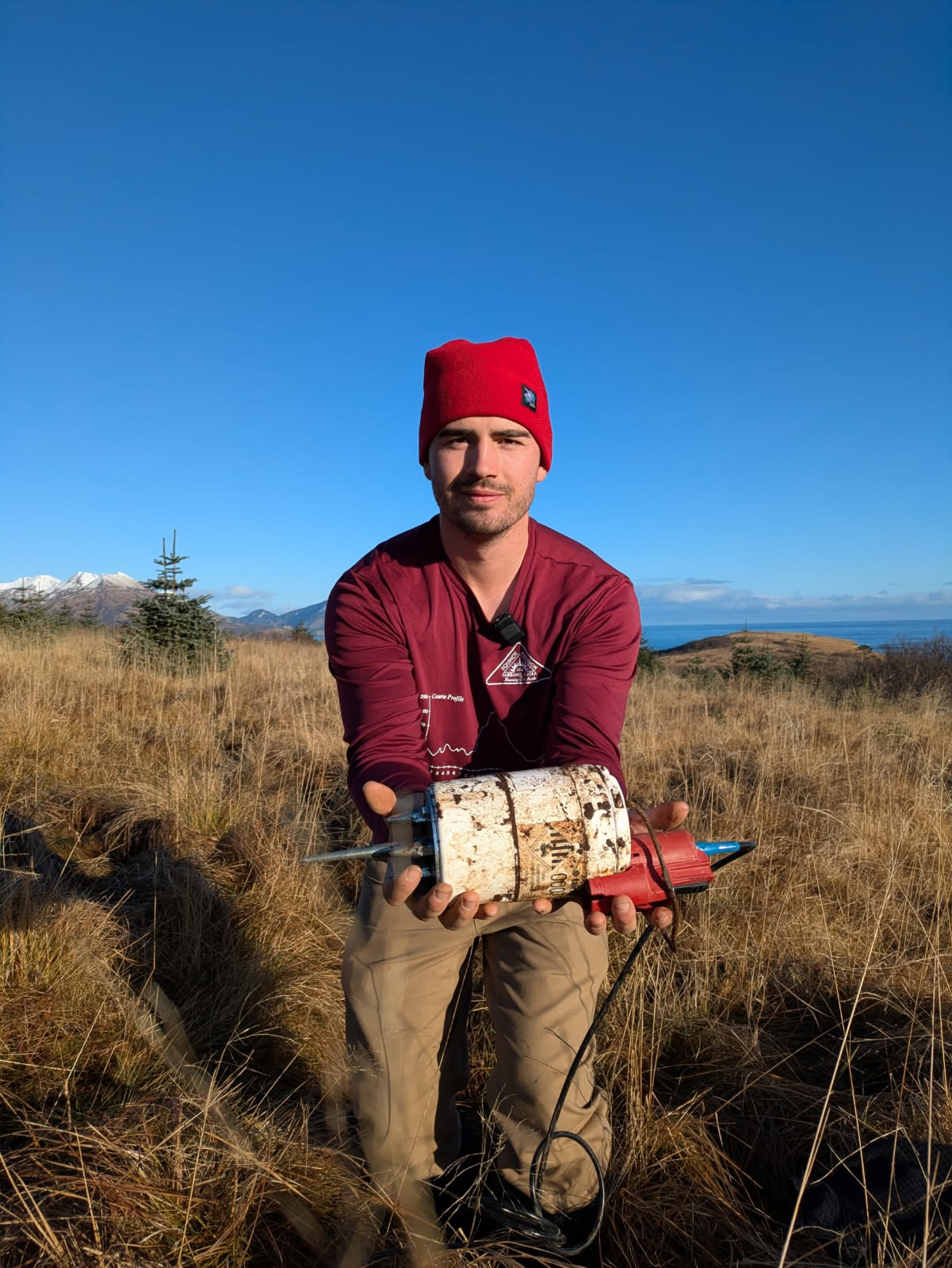 A man in a red knit cap, standing in a grassy fields with mountains and the ocean behind him, holds a dirty, white cylindrical piece of equipment, about a foot long, with an electrical cord hanging from it.