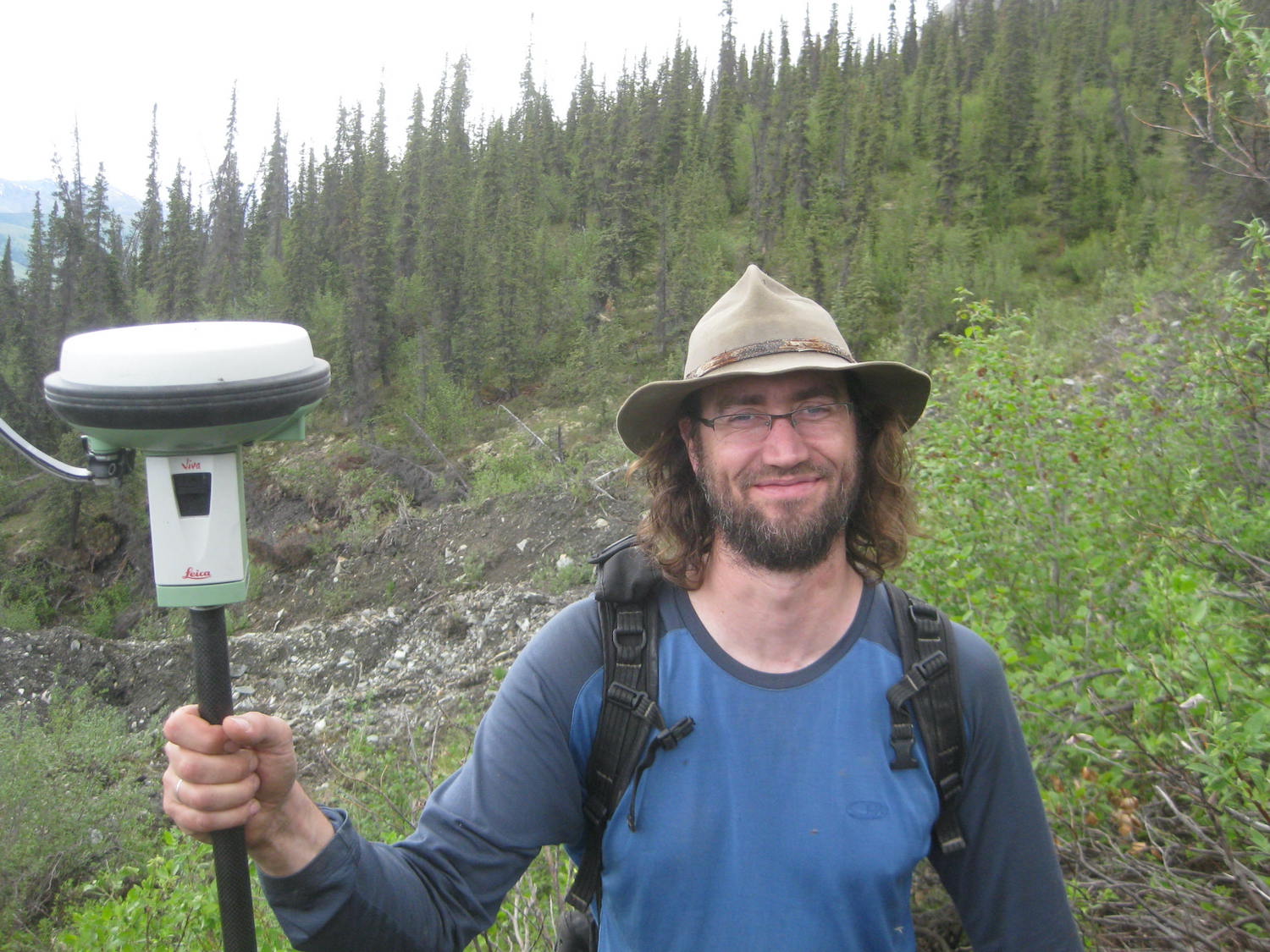 A bearded man with a wide-brimmed hat holds a satellite-based positioning receiver (a disc atop a staff) in front of a hillside covered mostly with spruce trees.