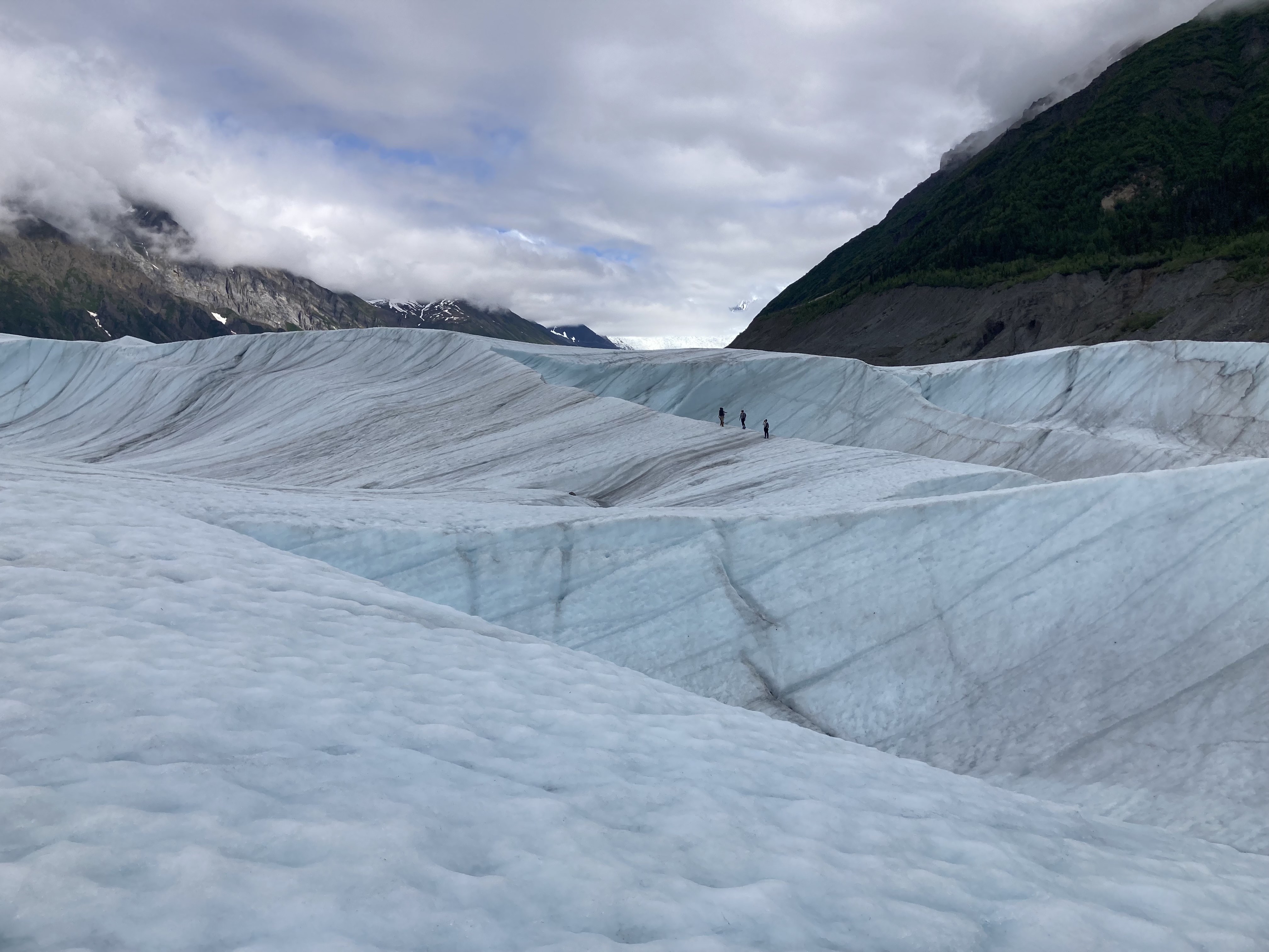 Participants in a 2022 international glaciology summer school walk across the rippled, blue-white surface of Root Glacier near McCarthy, Alaska. Three small figures move along a shallow ice ridge, surrounded by sweeping glacier valleys and steep, cloud-covered mountains in the distance.