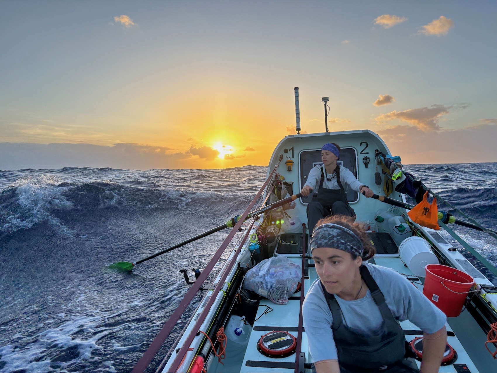 Two women row a boat with a cabin that sports buckets, water jugs and other items strapped to it. The boat rises on a wave while headed toward a golden sunset.