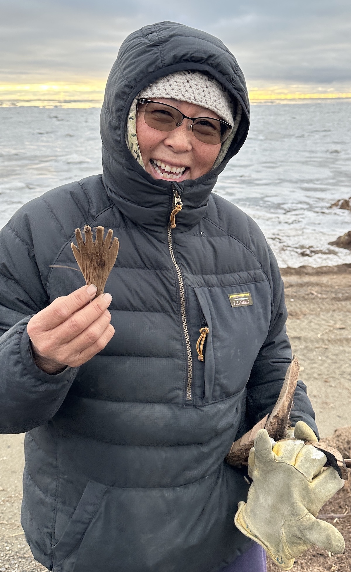 While standing on a beach, a woman in a black winter hooded coat holds a small hand-like figure carved from wood.