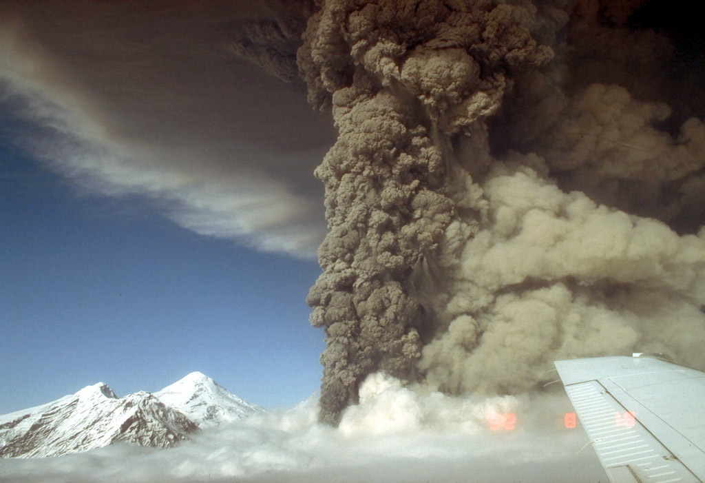 A towering dark gray ash plume rises from Mount Spurr’s Crater Peak vent during the Aug. 18, 1992, eruption. A lighter tan cloud from pyroclastic flows spreads to the right, while the snow-covered 11,070-foot summit lava dome complex is visible at left beneath a blue sky. Part of an aircraft wing appears in the lower right.
