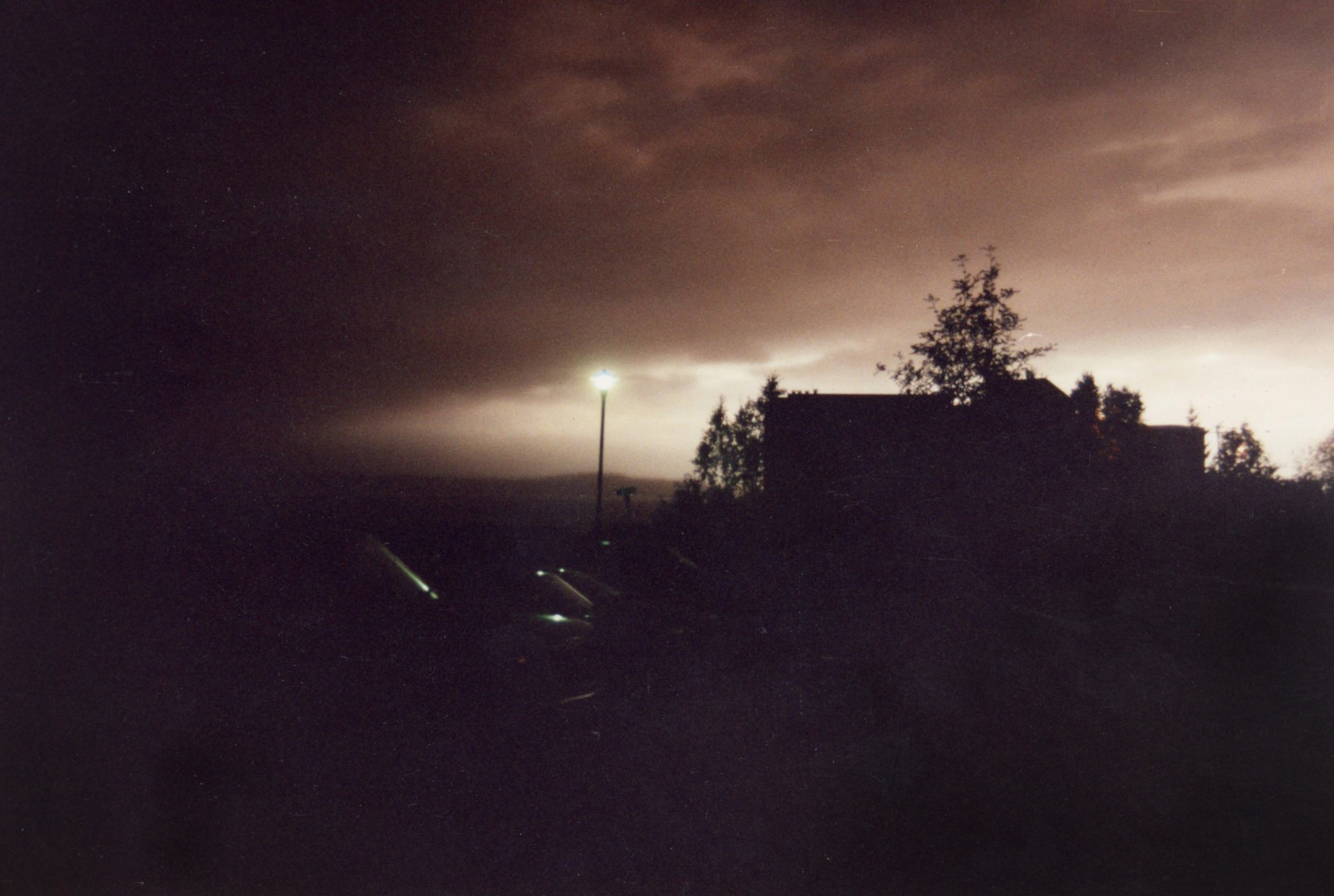 A dark brown-gray ash cloud from Mount Spurr spreads over Anchorage on Aug. 18, 1992, dimming the late-afternoon sky above Rod Boyce’s condominium near Cook Inlet. A bright band remains near the horizon while buildings, trees and parked cars appear in silhouette and a streetlight glows beneath the advancing cloud.