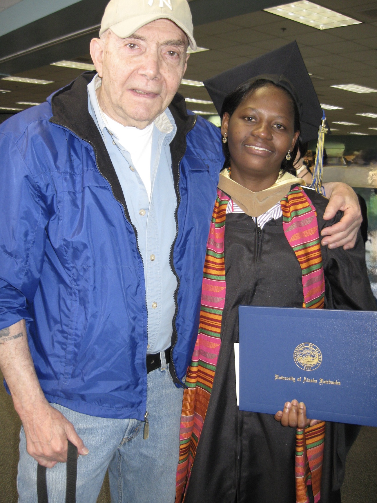  An older man stands next to a young woman dressed in academic regalia and holding a diploma. 
