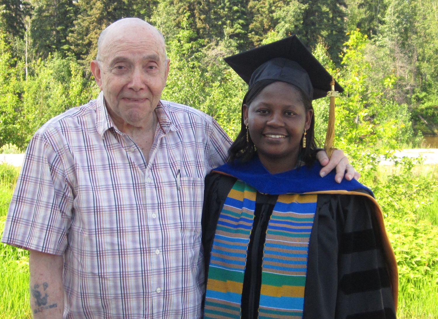 An older man stands outside next to a young woman dressed in academic regalia. In the background, sun brightens green vegetation. 