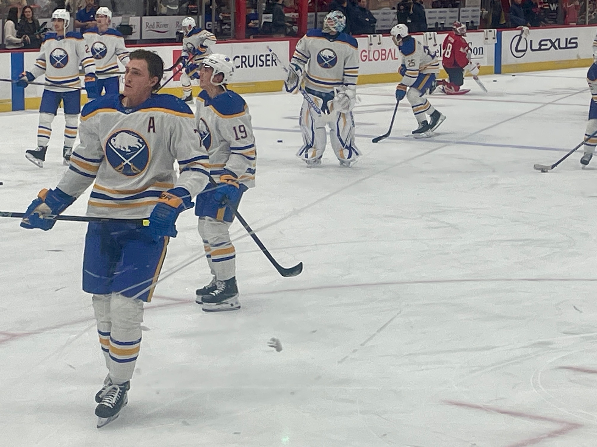 Hockey players dressed in white, blue and gold uniforms glide across an ice rink. The one closest to the camera has no helmet.