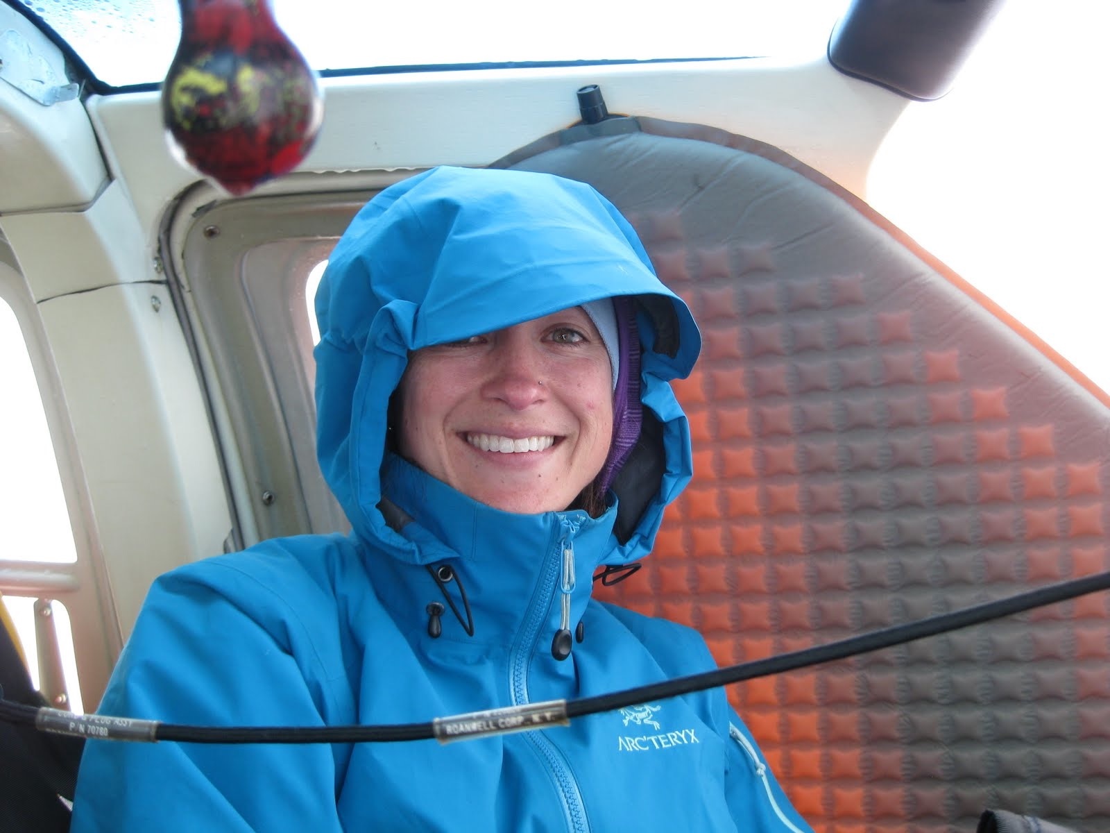 A woman in a blue hooded raincoat smiles. A sleeping pad rests against the inside of a helicopter window behind her.