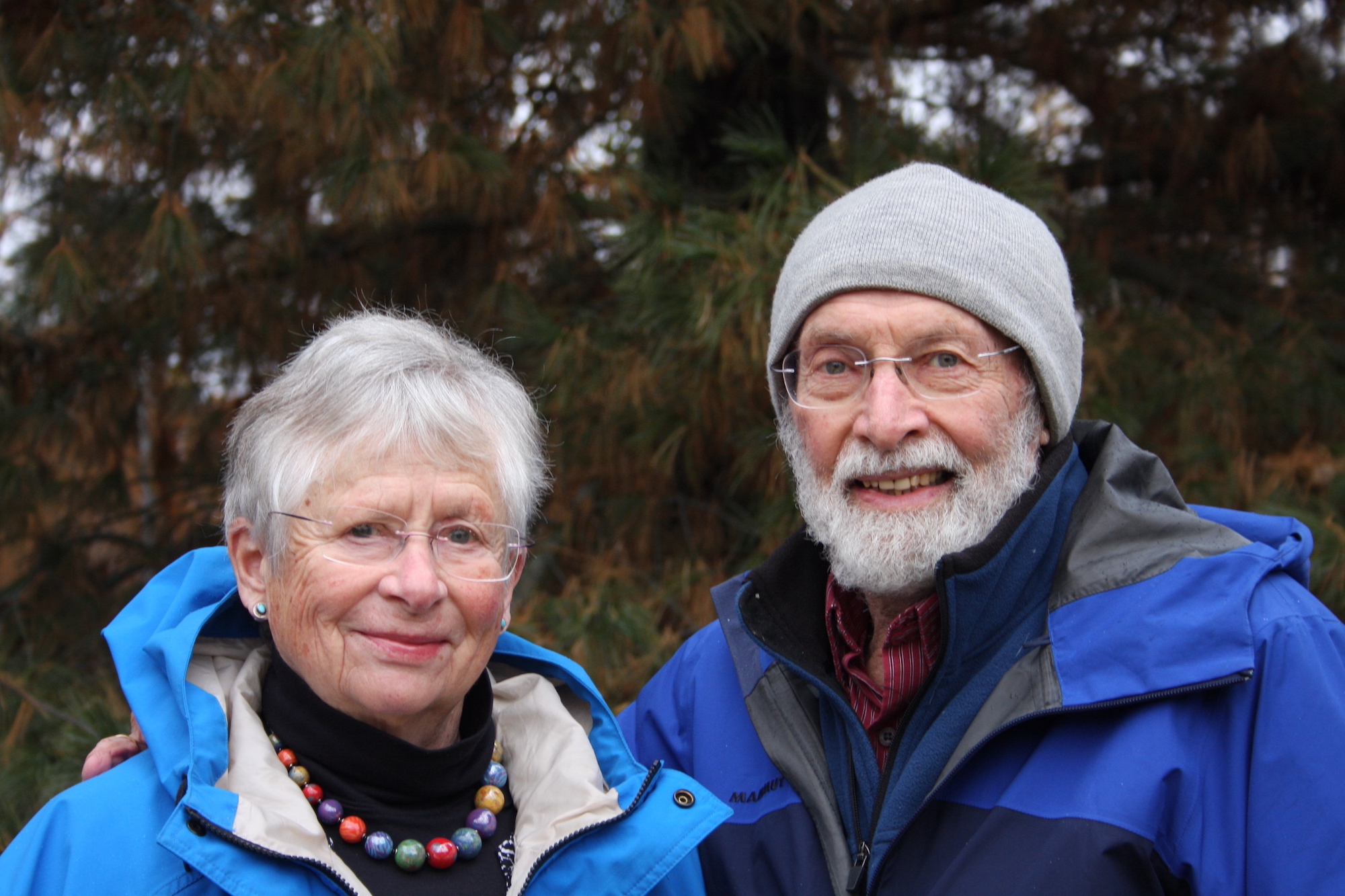 A woman and a man, both with gray hair, glasses and wearing blue raincoats, smile at the photographer. The man has a beard and a knit cap.