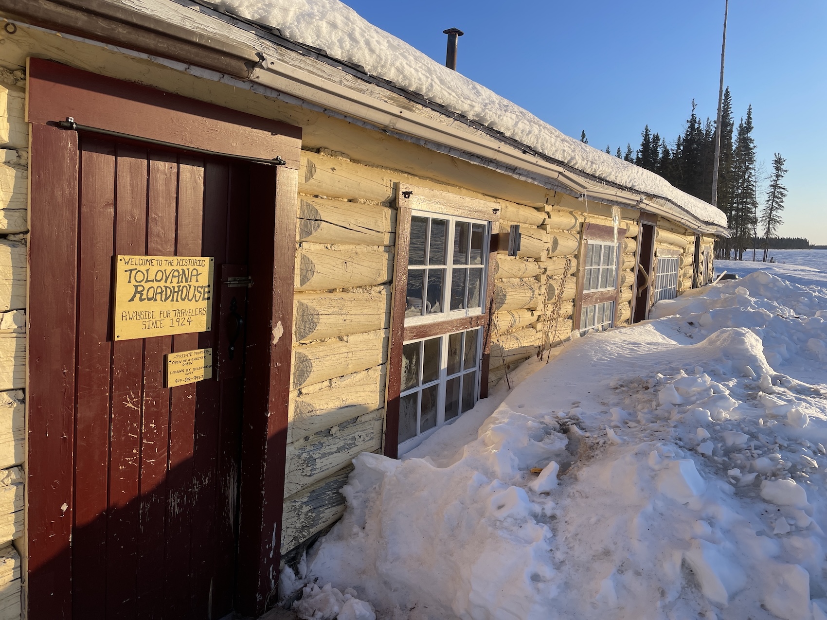 A long, one-story log cabin sits in low sunlight under a blue sky, with spruce trees rising on the bank of a frozen river in the background. The cabin logs are painted yellow. On the painted red door, a sign says "Welcome to the historic Tolovana Roadhouse, a wayside for travelers since 1924. 