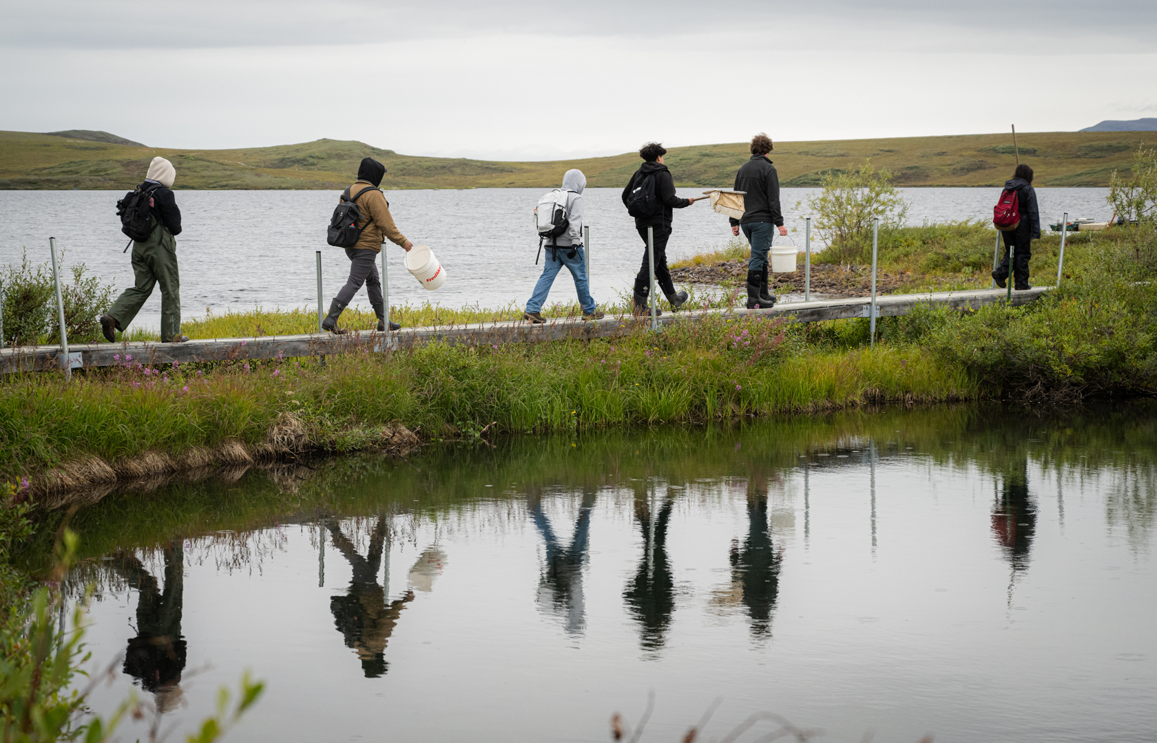 A group of students walk across a bridge at Toolik Field Station with their reflections in the pond below them.