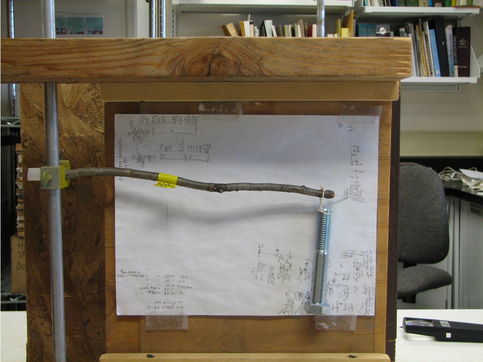 A stick mounted horizontally, with a weight hanging off the end, in front of a piece of paper mounted on plywood. 
