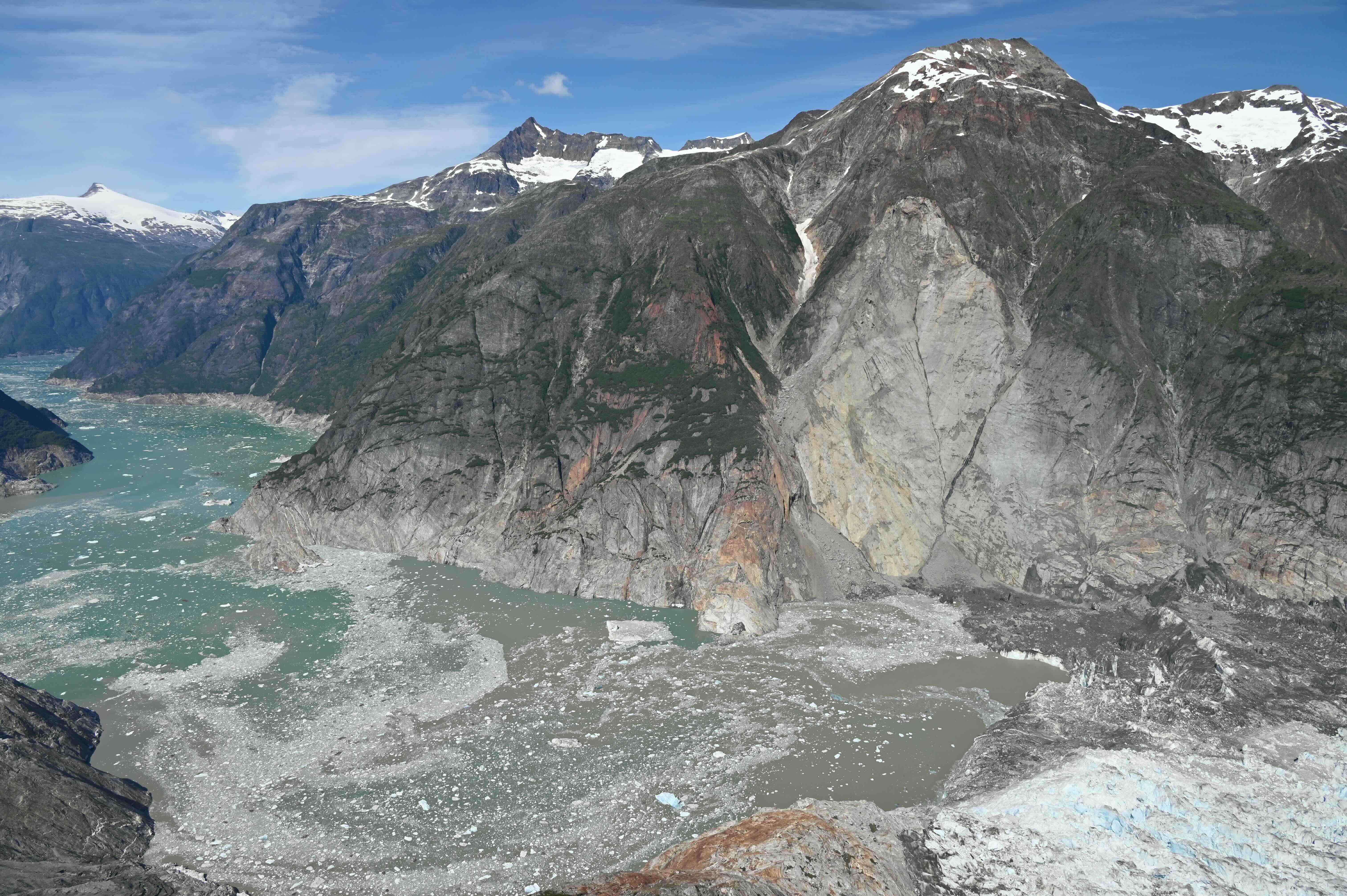 A wide aerial view of Tracy Arm in Southeast Alaska shows the scar from the Aug. 10, 2025, landslide descending from a steep mountainside into the fjord near a glacier terminus. Gray rock and debris cut through the center of the mountain, reaching the water below, where floating ice and sediment spread across the fjord. The glacier terminus is visible at lower right, with snow-covered peaks rising above the surrounding mountains. Along the far side of the fjord, a pale trimline marks where the tsunami stripped vegetation from the slopes. The photograph was taken from across the fjord during a U.S. Geological Survey flight on Aug. 13, 2025.