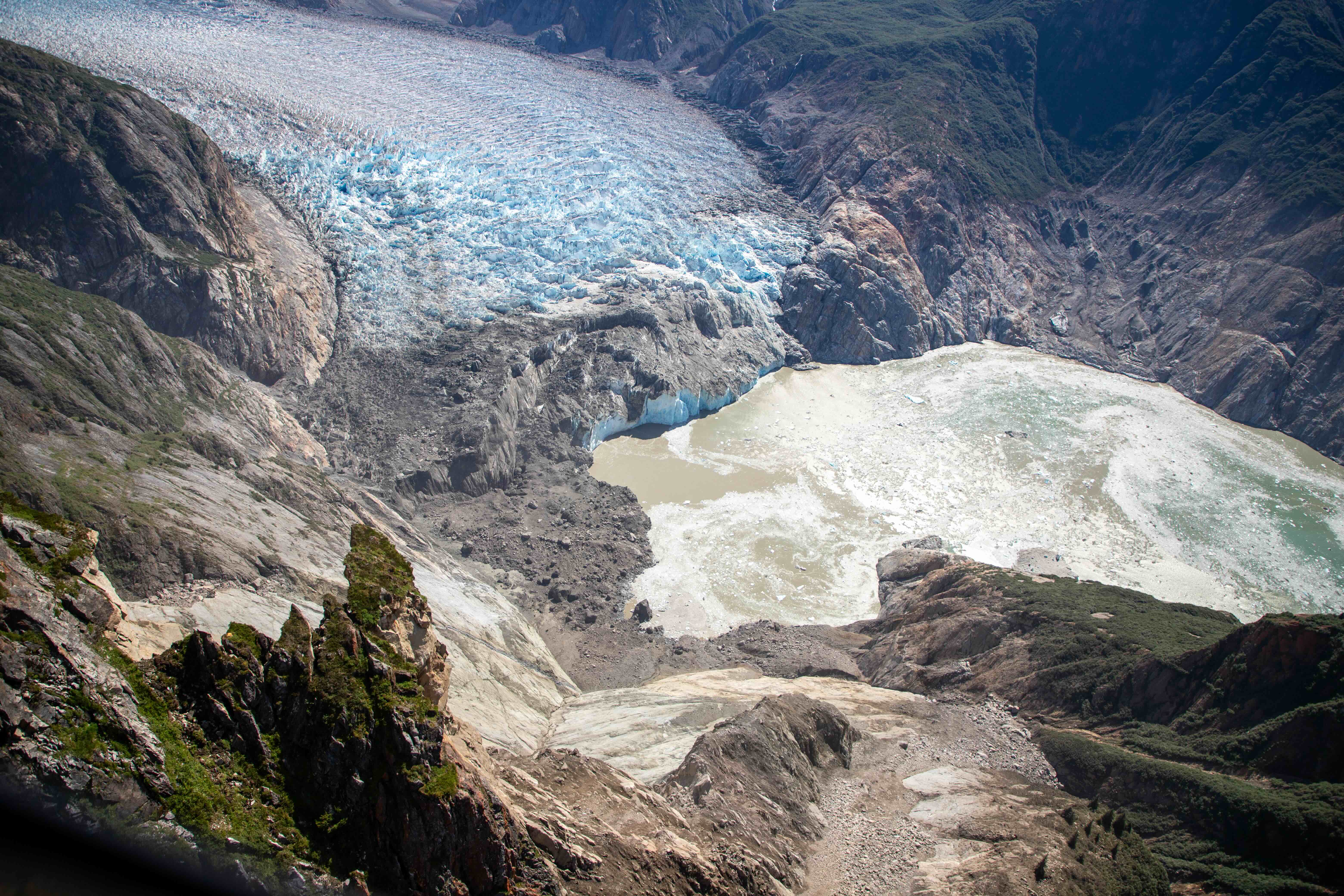 An aerial photograph looks down into the steep-walled Tracy Arm fjord in Southeast Alaska. In the foreground, a broad landslide scar cuts down a rocky mountainside toward the water, leaving exposed gray and tan rock and scattered debris. Across the water, the opposite slope shows the area of highest tsunami run-up from the landslide-triggered wave, where vegetation was stripped away along the fjord wall. A glacier terminus with blue ice and dark sediment sits at the head of the inlet beyond the water. The photograph was taken Aug. 13, 2025, during a U.S. Geological Survey field reconnaissance overflight following the Aug. 10, 2025, landslide and tsunami.