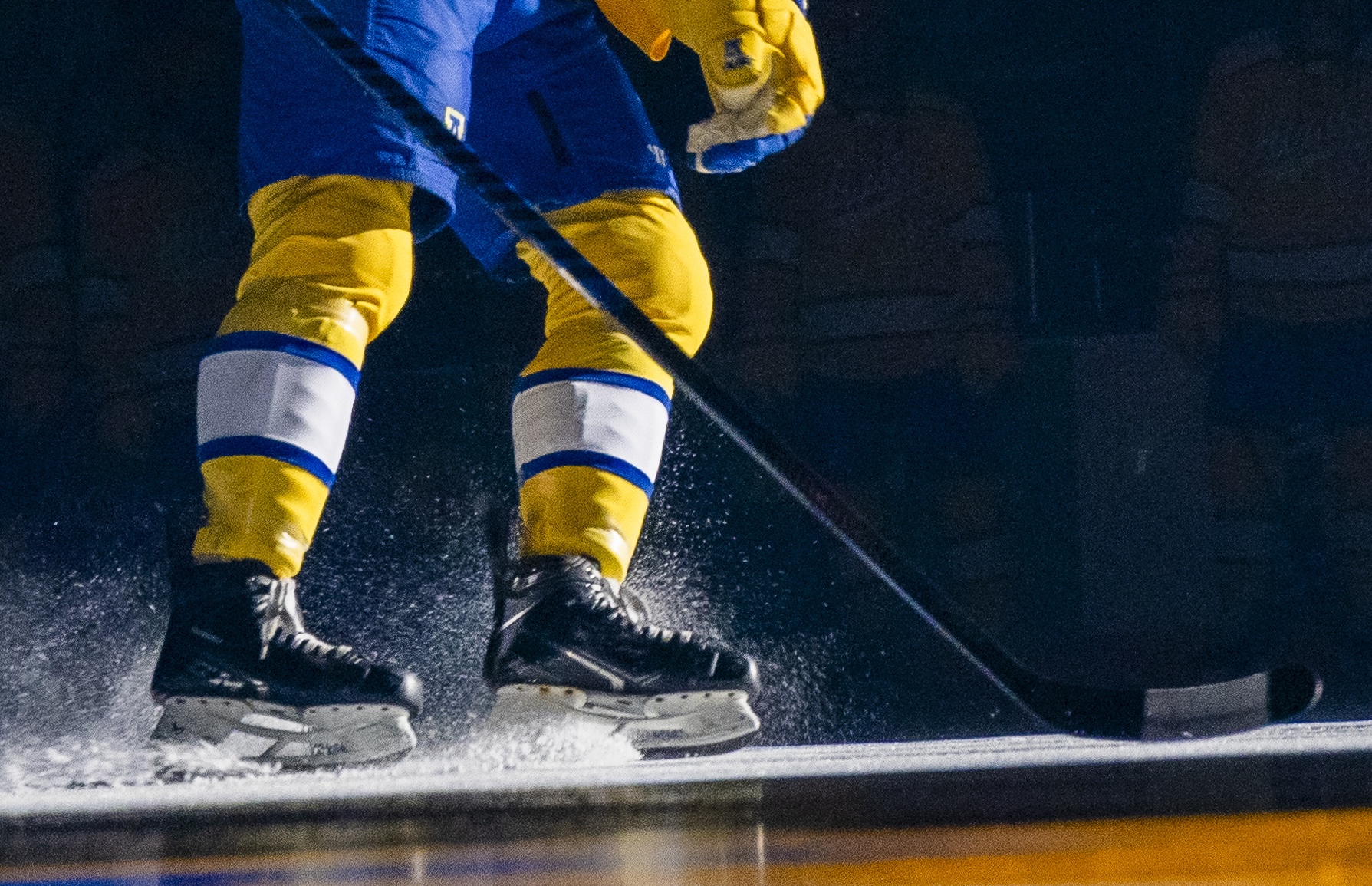 A close-up photo captures shavings spraying from skate blades as a hockey player's legs, dressed in blue and gold, turn on an ice rink surface. The blade of a hockey stick slides on the rink in front of the ice skates. 