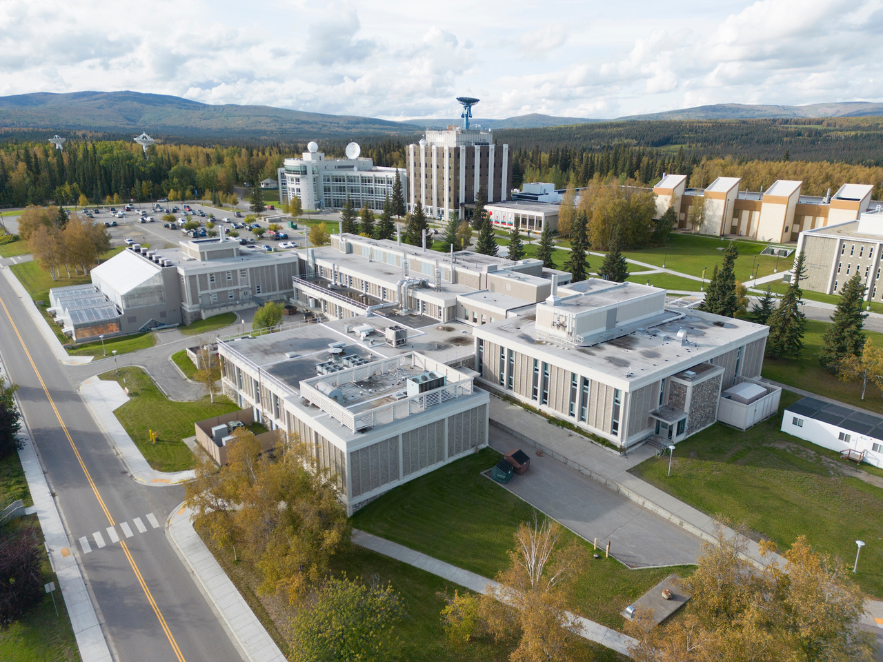 Aerial view of the University of Alaska Fairbanks Arctic Research Park, showing research and office buildings surrounded by roads, lawns and autumn trees, with forested hills in the distance under a partly cloudy sky.