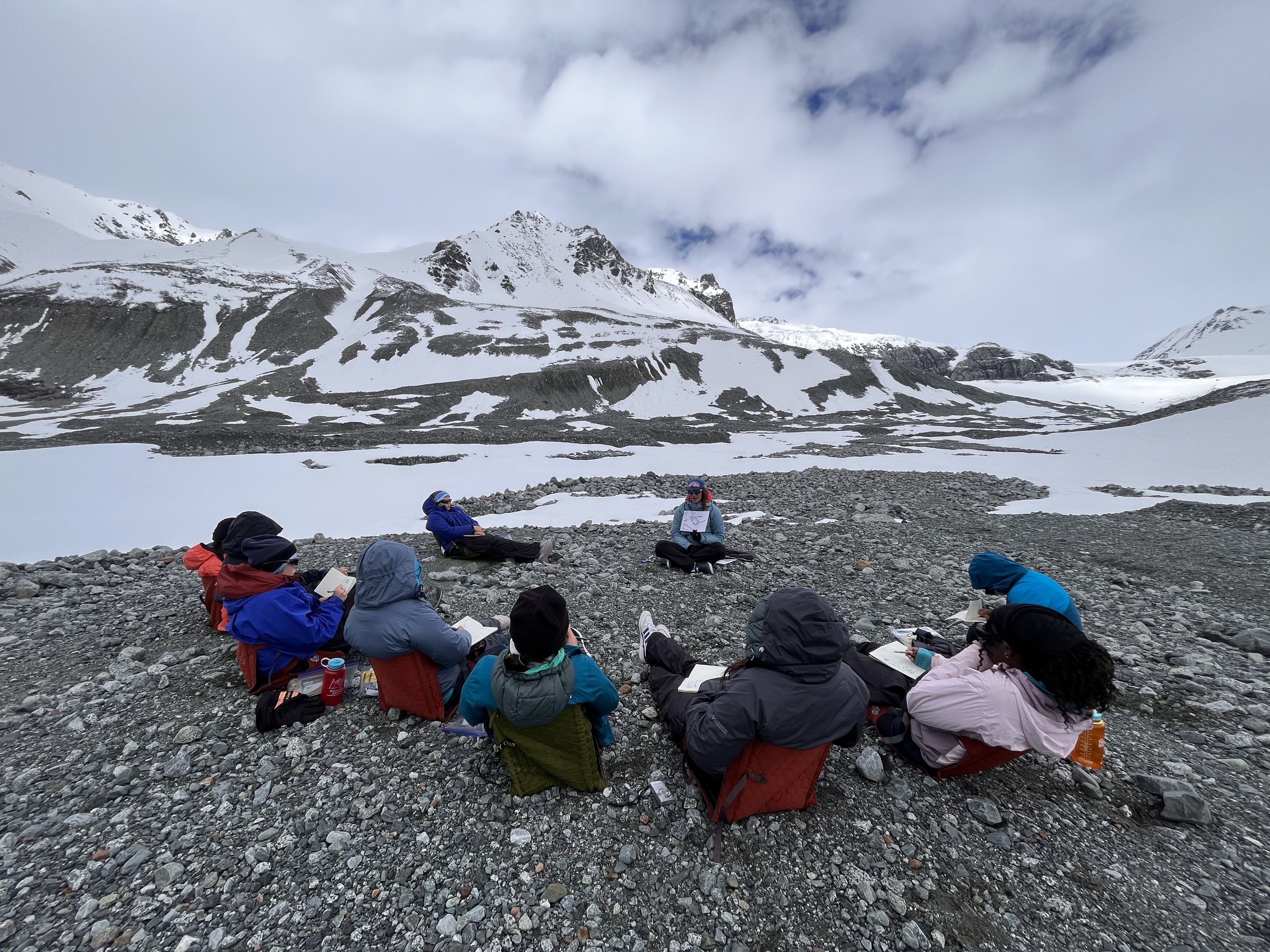 A group of young women sit in a circle in front of Gulkana Glacier learning from an instructor.