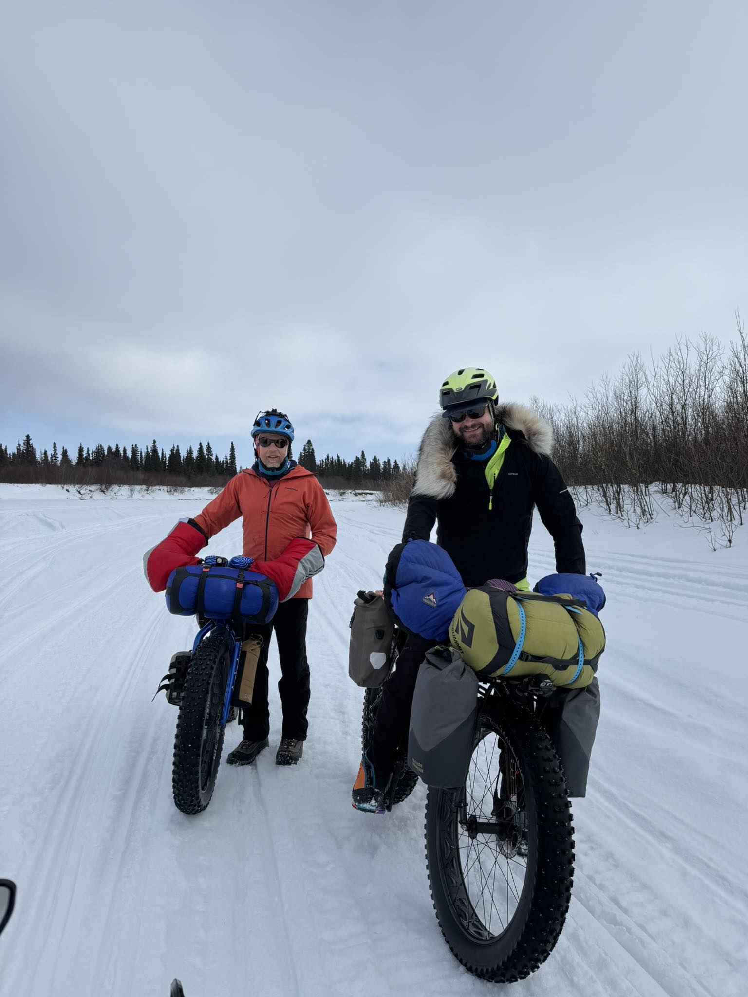 Two men stand next to fat-tired bikes laden with outdoor gear. They're on a snowmachine trail on a river. In the background are willows and spruce along the riverbanks.