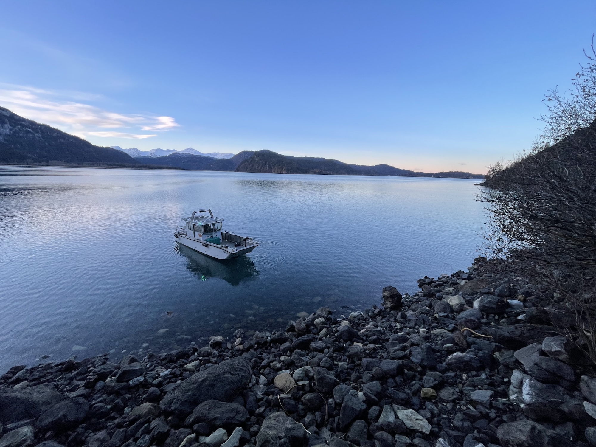 A boat rests on calm water near a rocky beach. In the background are sunlit mountains.