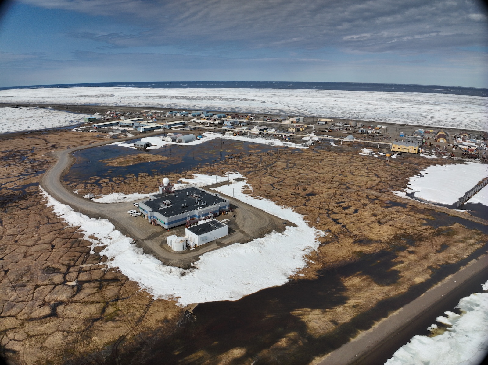 A aerial view shows a large building sitting on a gravel pad ringed by snow drifts in an otherwise bare spot of grassy tundra. On the other side of the building are a few streets lined with buildings along a gravel beach coastline, beyond which is an expanse of sea ice and then the open ocean.