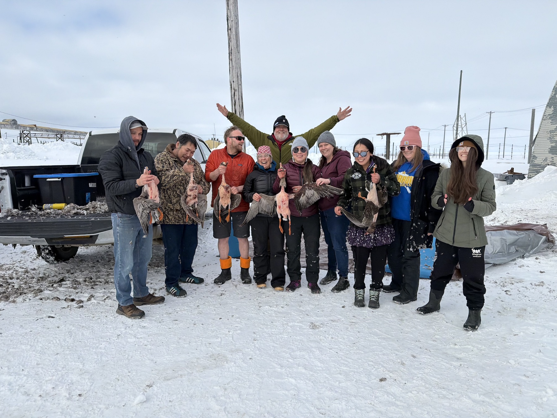 Ten men and women wearing warm coats and hats stand on snowy ground while holding several plucked geese.